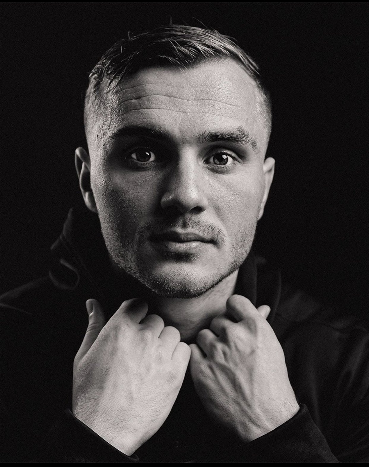 Black and white portrait of a man with short hair, looking directly at the camera, with hands touching his neck.