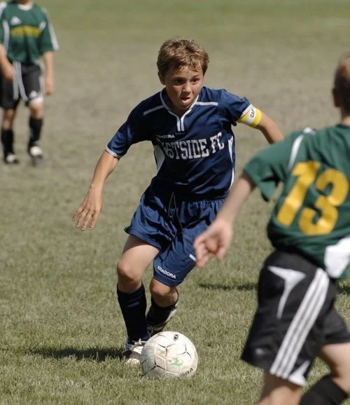 Young boys playing soccer on a grassy field, one in a blue uniform dribbling the ball while others in green jerseys play nearby.