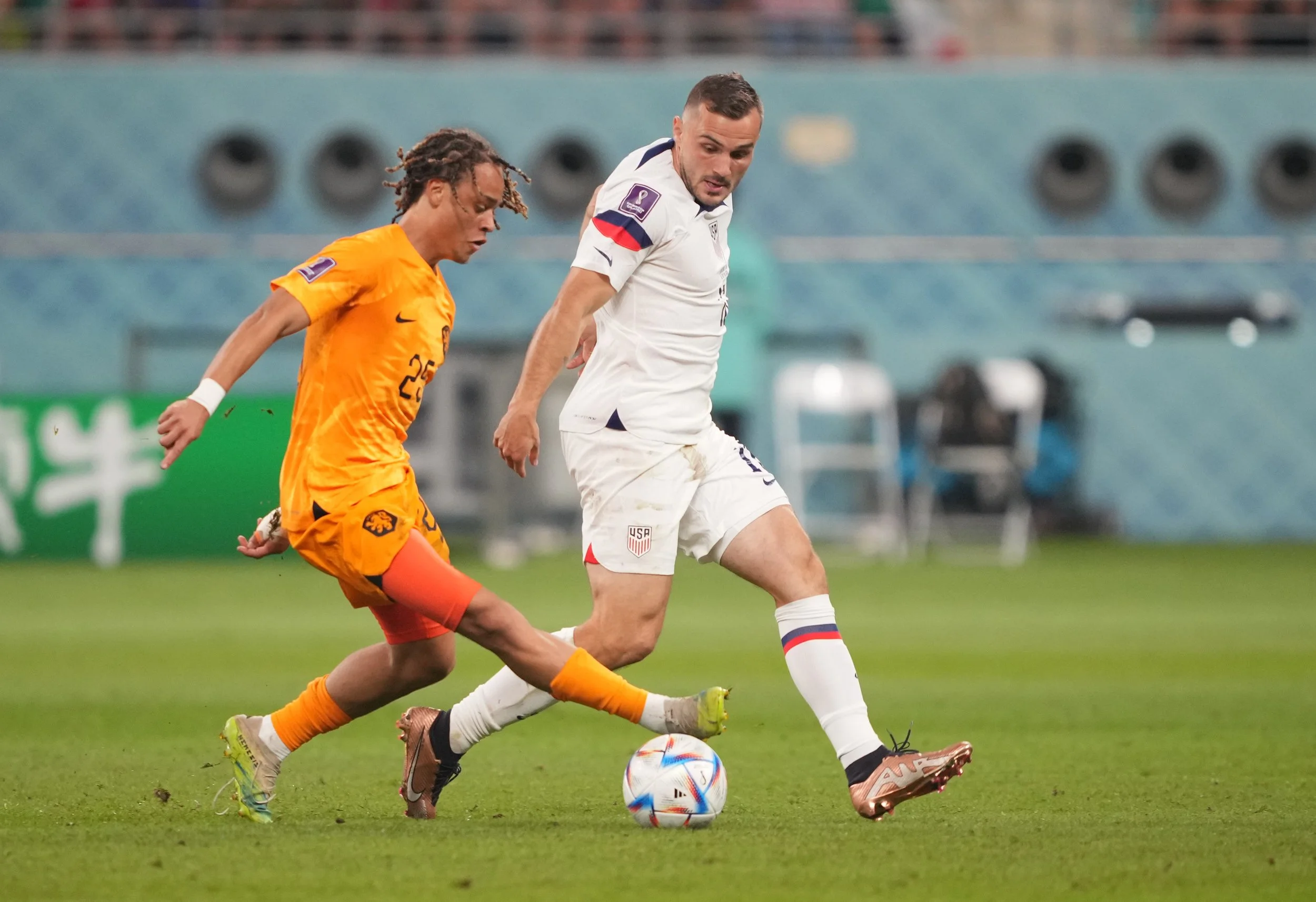 Two soccer players, one in an orange jersey and the other in a white jersey, compete for the ball on the field.