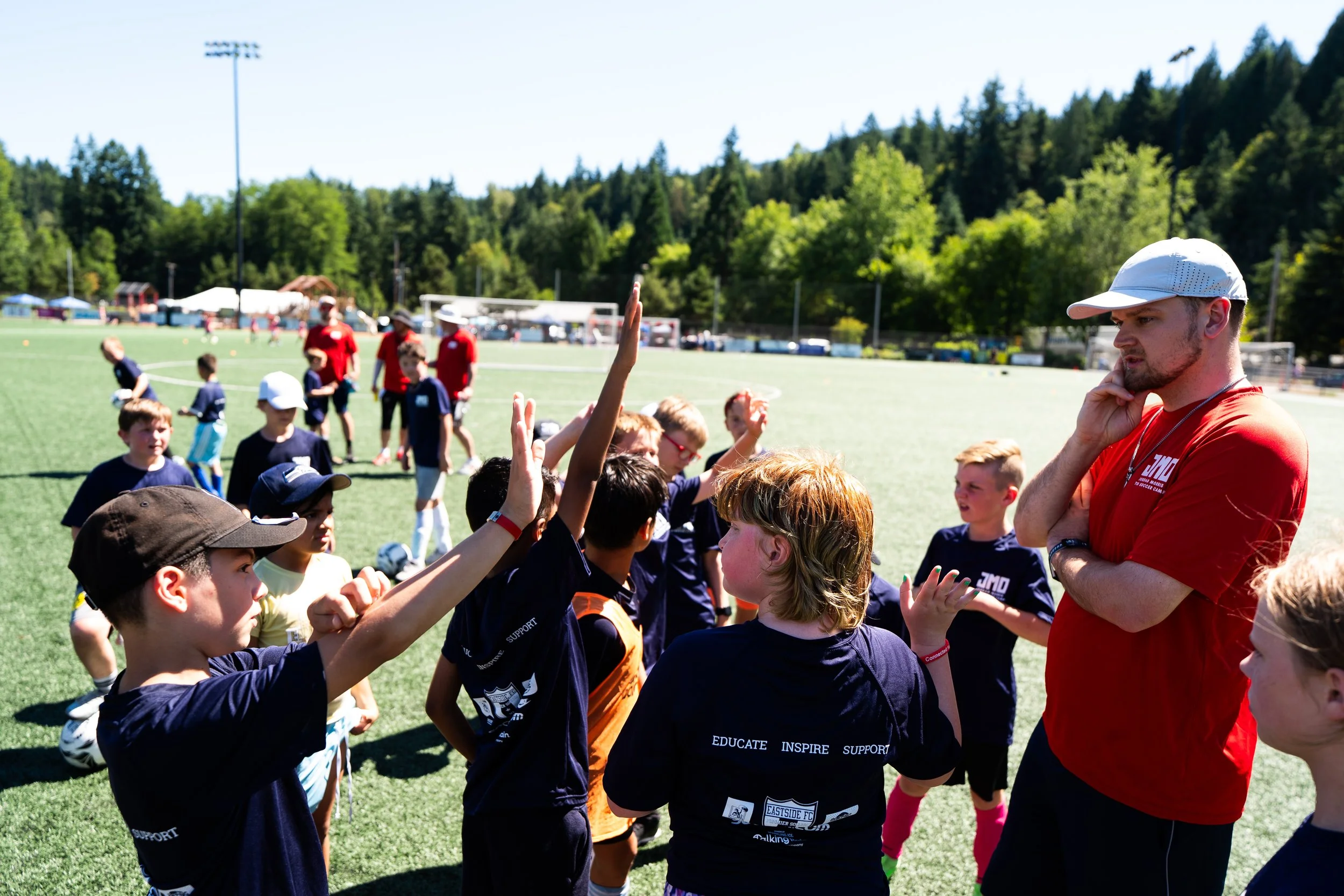 Group of children and adult coaches on a soccer field with a background of trees and a mountain, during what appears to be a youth soccer event.
