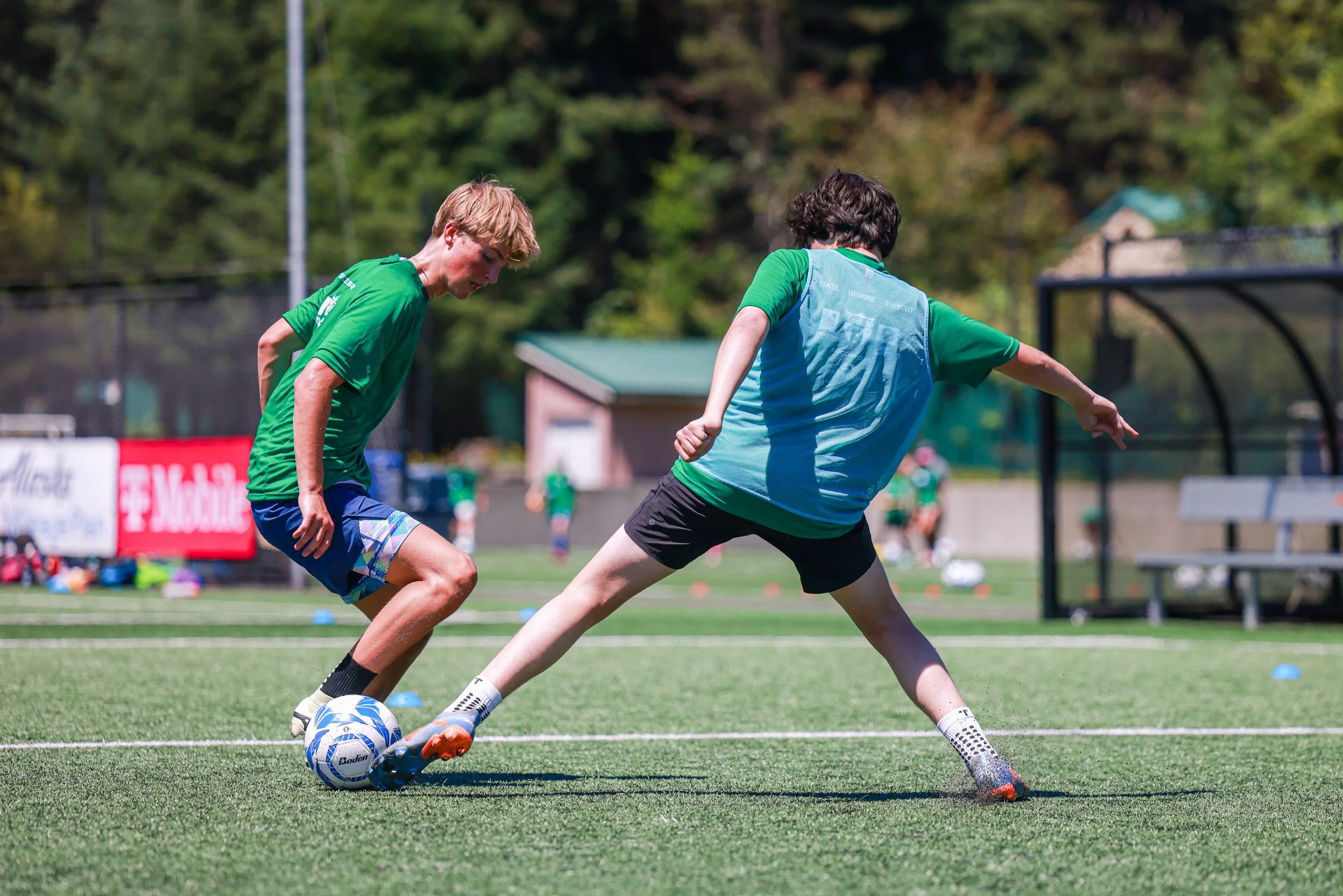 Two boys playing soccer on a field, one in a green shirt and shorts and the other in a green and blue shirt, both trying to gain control of the ball.