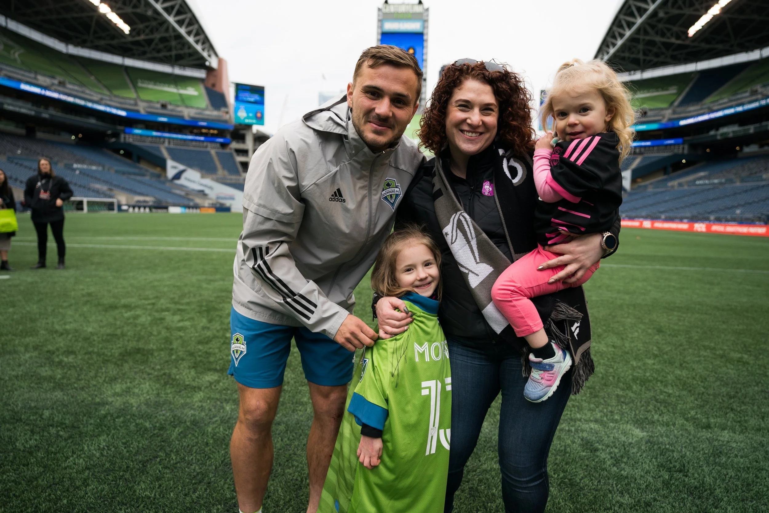 A family of four, including two children, taking a photo together on a soccer field inside a stadium. The adults are smiling, and the children are dressed casually. The stadium seats and large screens are visible in the background.