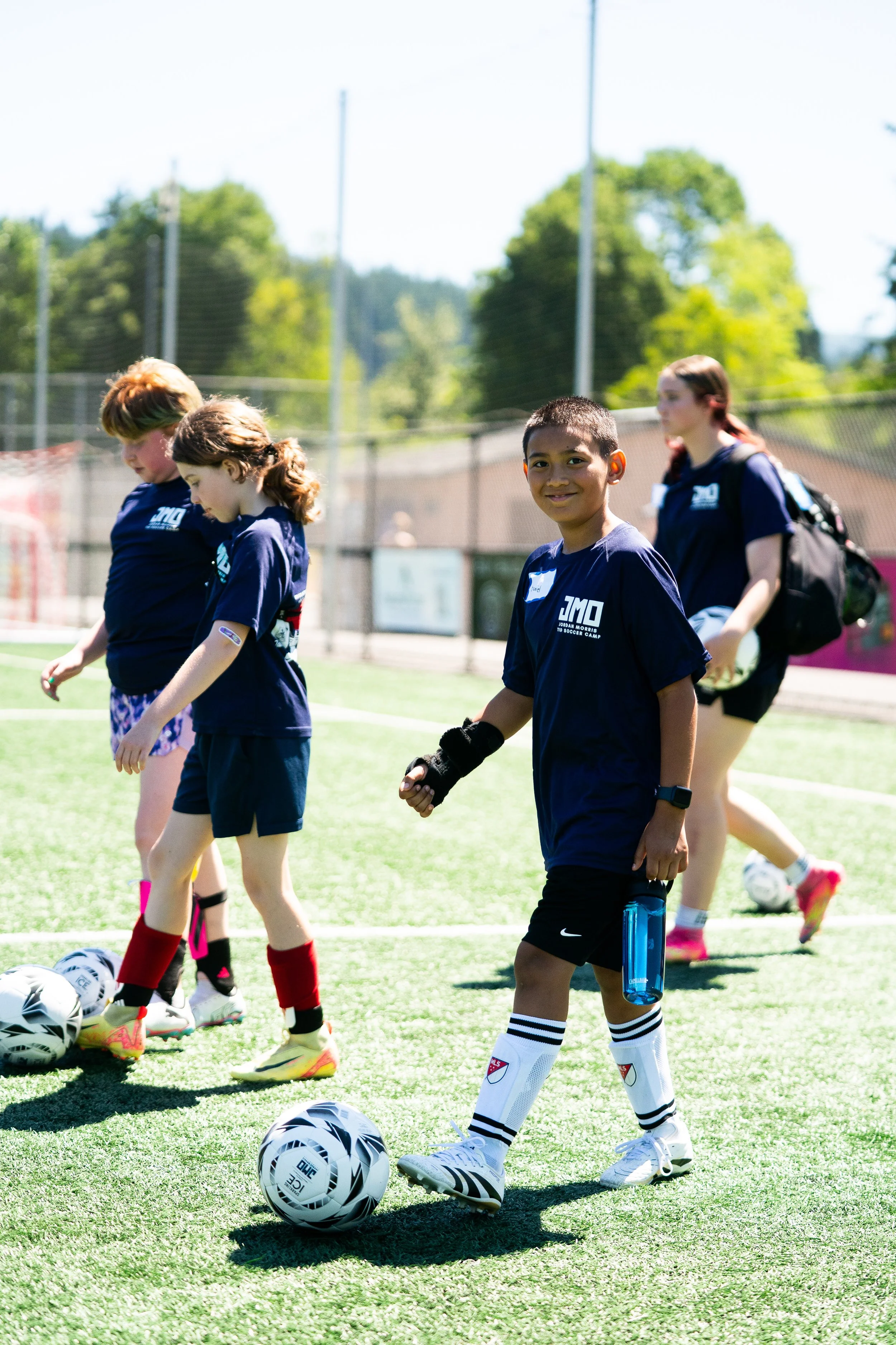 Children in soccer uniforms on a soccer field during practice, with soccer balls on the ground.