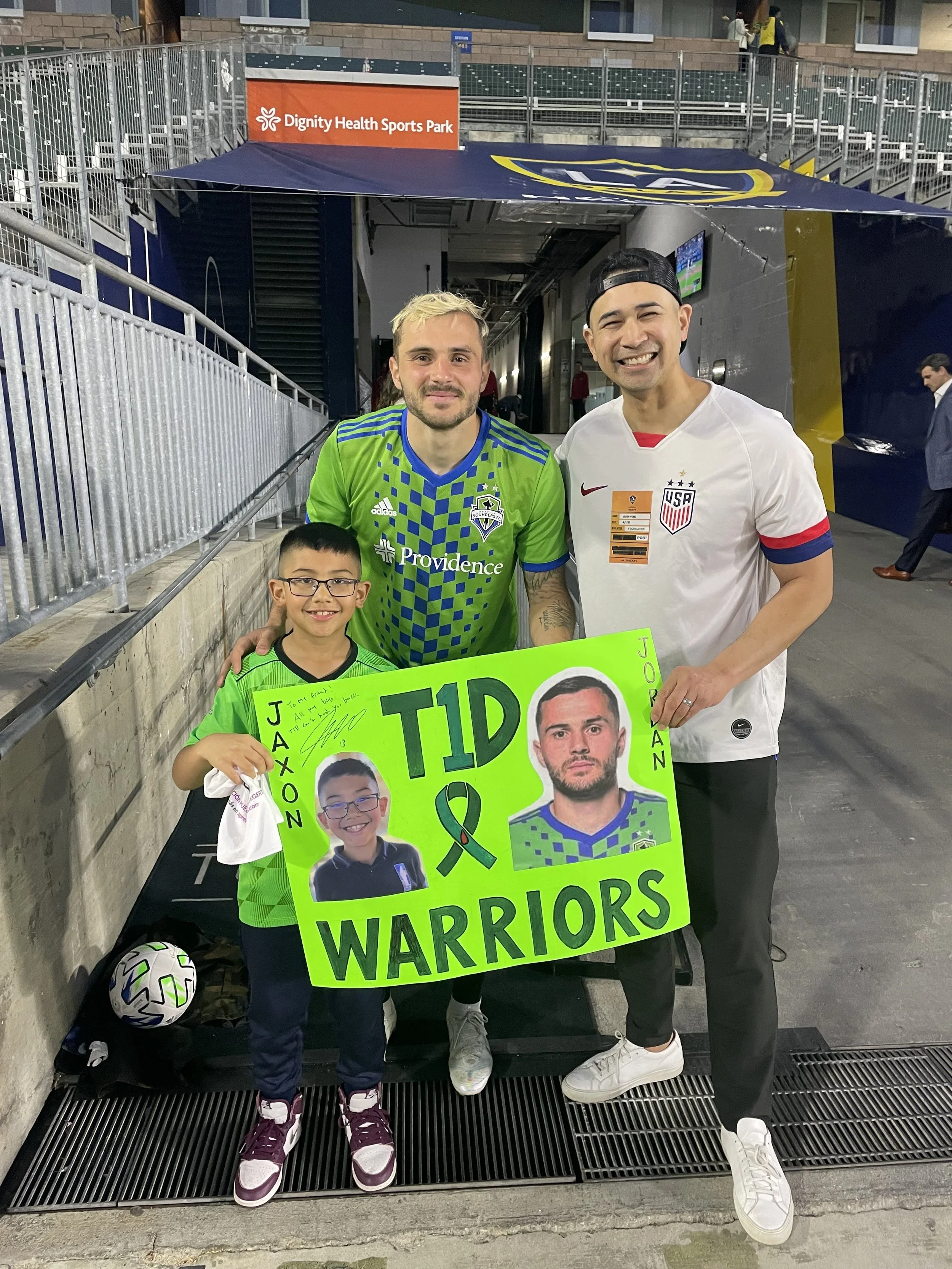 Three people, two adults and one child, standing together at a sporting event, holding a bright green sign that reads 'TID of WARRIORS' with photos of two young athletes. The adults are wearing soccer jerseys, one with a Seattle Sounders FC kit and the other wearing an USA national team shirt. The child is in an athletic outfit, holding a soccer ball, and wearing glasses.