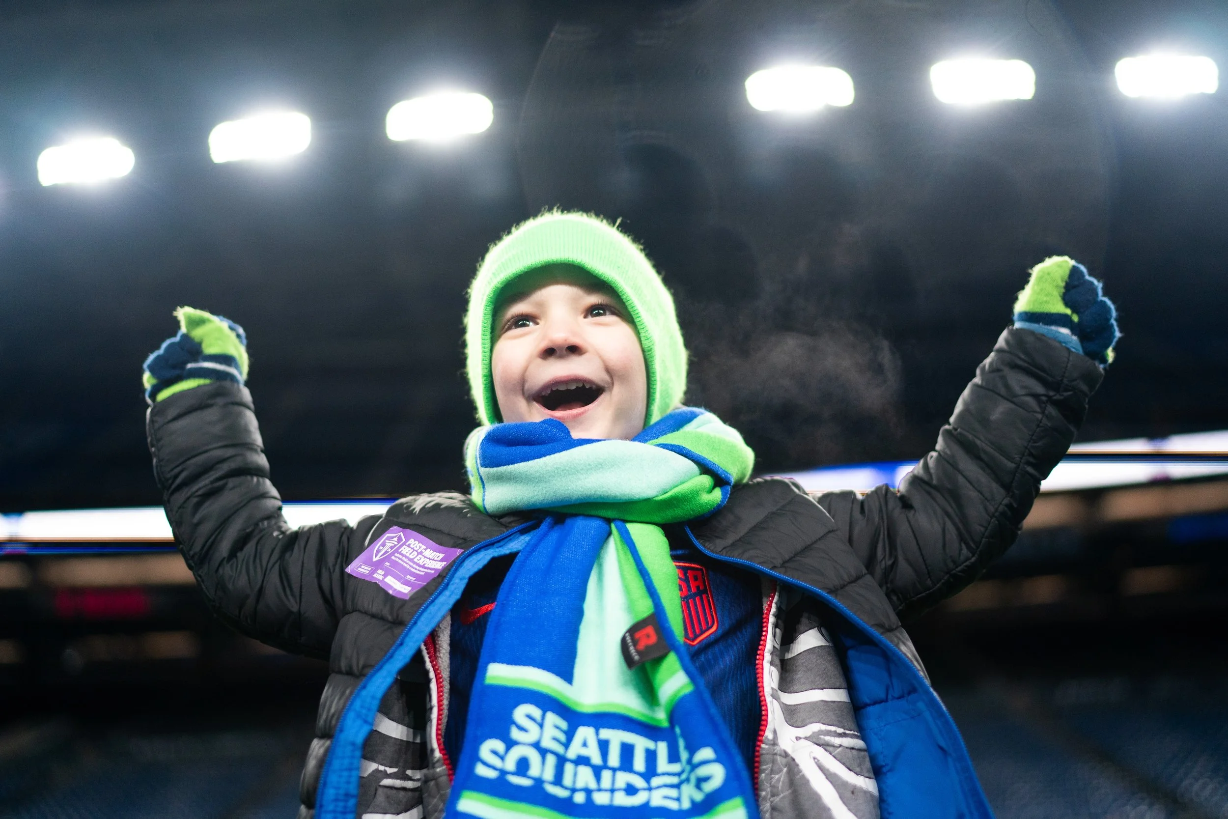 A young boy wearing a green knit hat, a colorful scarf, and a black jacket with a Seattle Sounders patch, smiling and raising his arms in excitement at a stadium with bright overhead lights.