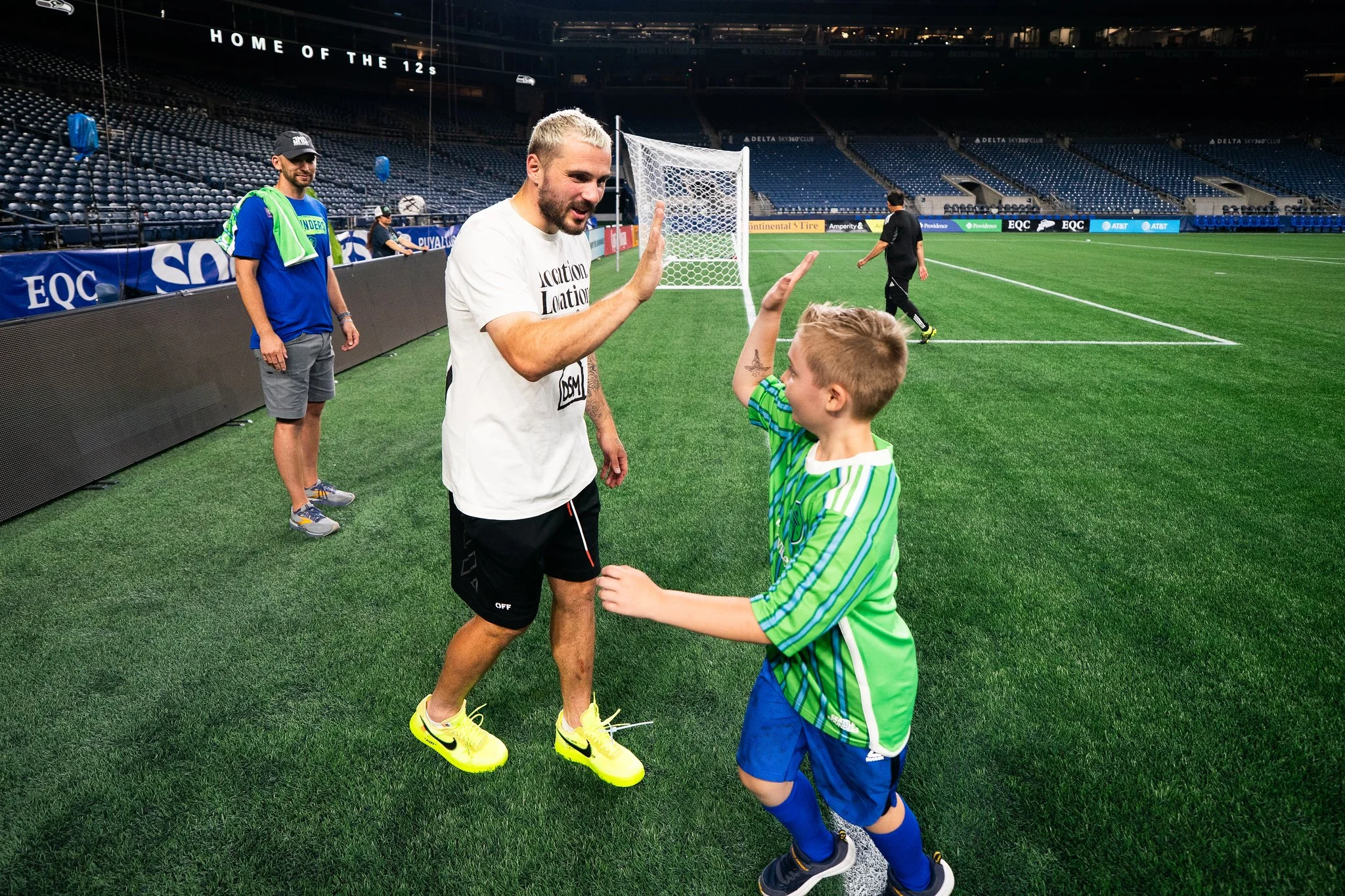 A man in a white shirt and black shorts high-fives a young boy in a green and blue sports jersey on the soccer field. Another man in a blue shirt and cap watches in the background, standing near the sideline of the stadium. The stadium is mostly empt