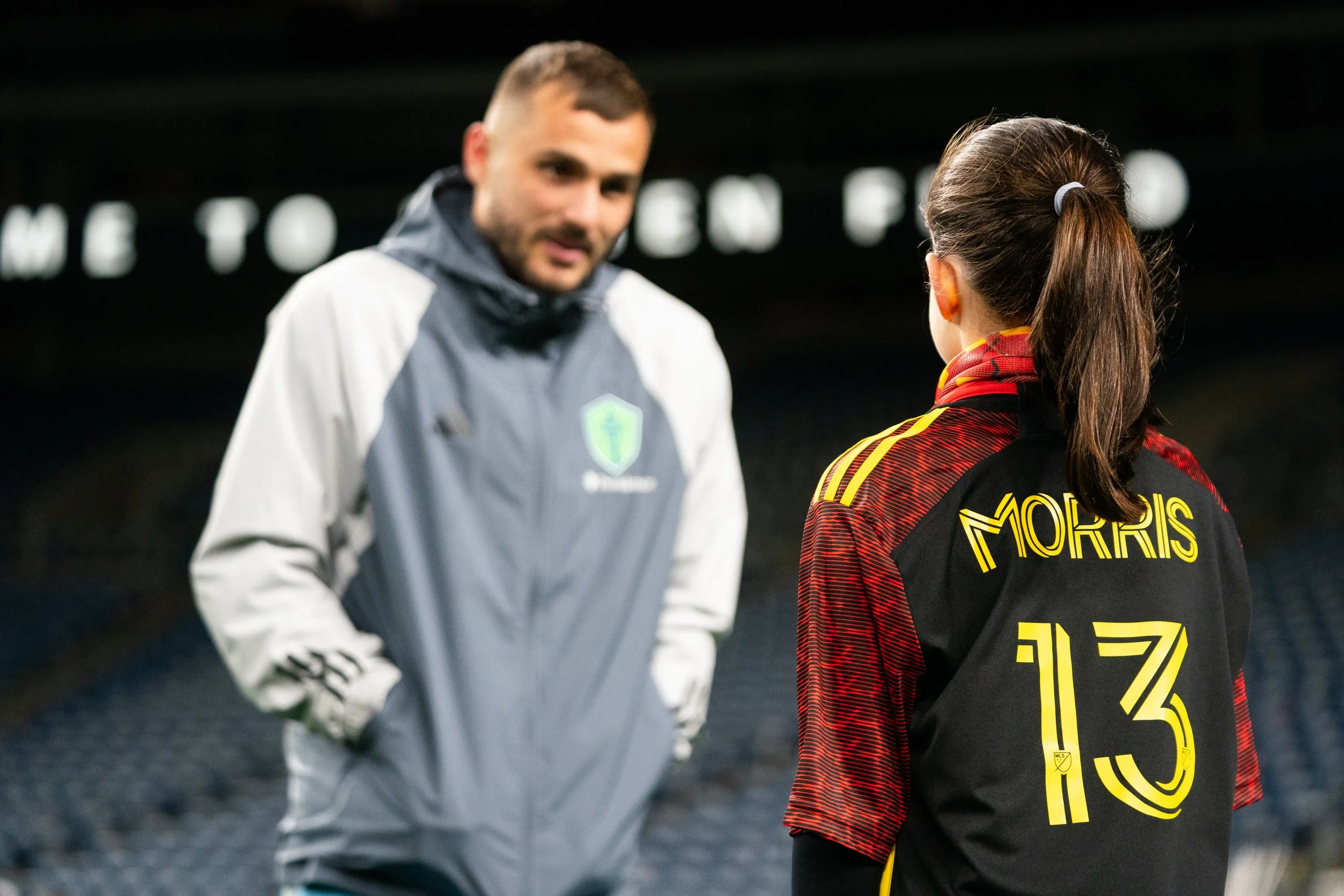 Two people talking outdoors at night, one wearing a gray sports jacket and the other in a black and red sports jersey with yellow lettering.