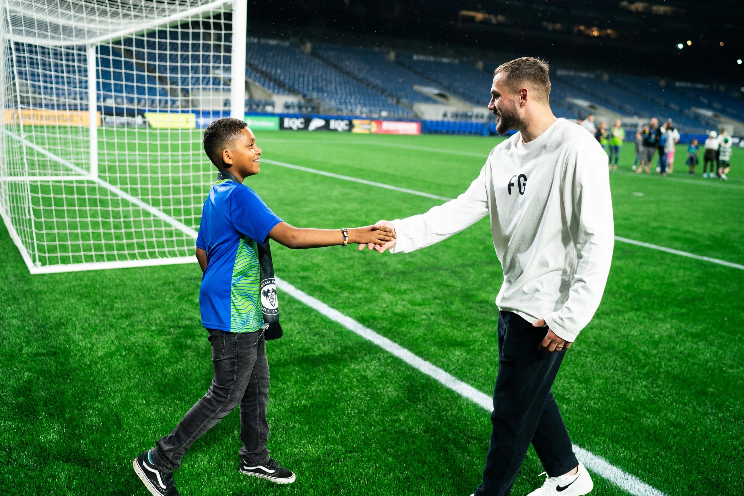 A young boy and a man shake hands on a soccer field at night, near the goalpost. The boy is wearing a blue sports jersey, and the man is dressed in a white sweatshirt with initials. In the background, there are spectators and other people on the fiel