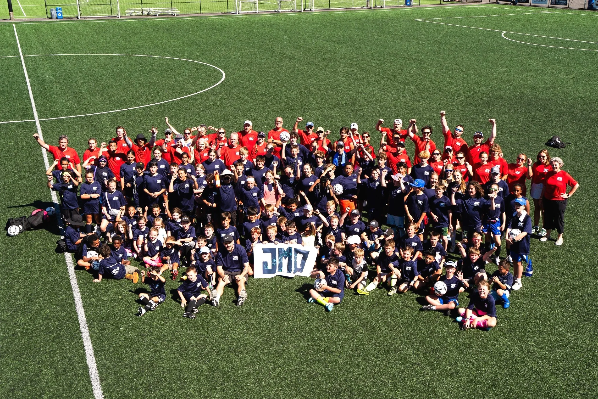 A large group of children and adults gathered on a soccer field, posing for a group photo. The group includes children in navy blue shirts and adults in red shirts, with some individuals holding soccer balls. A sign with the letters 'JMC' is visible in front of the group.