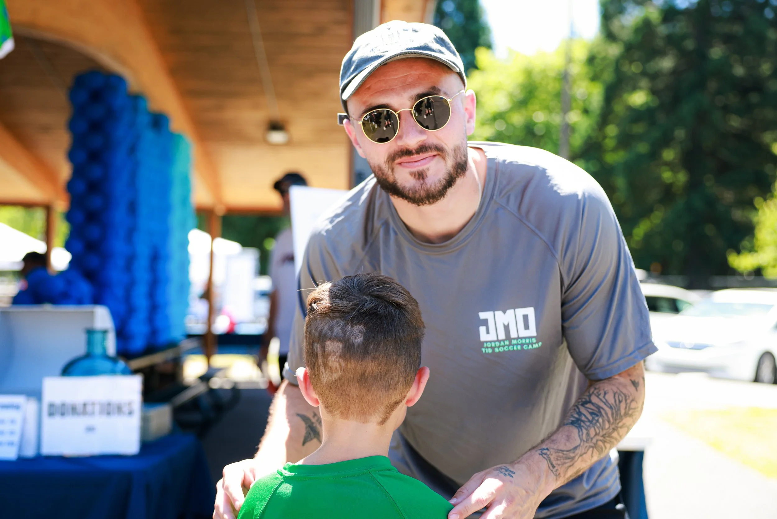 A man with tattoos and wearing sunglasses is smiling and talking to a young boy in a green shirt at an outdoor event with a food or merchandise stand in the background.