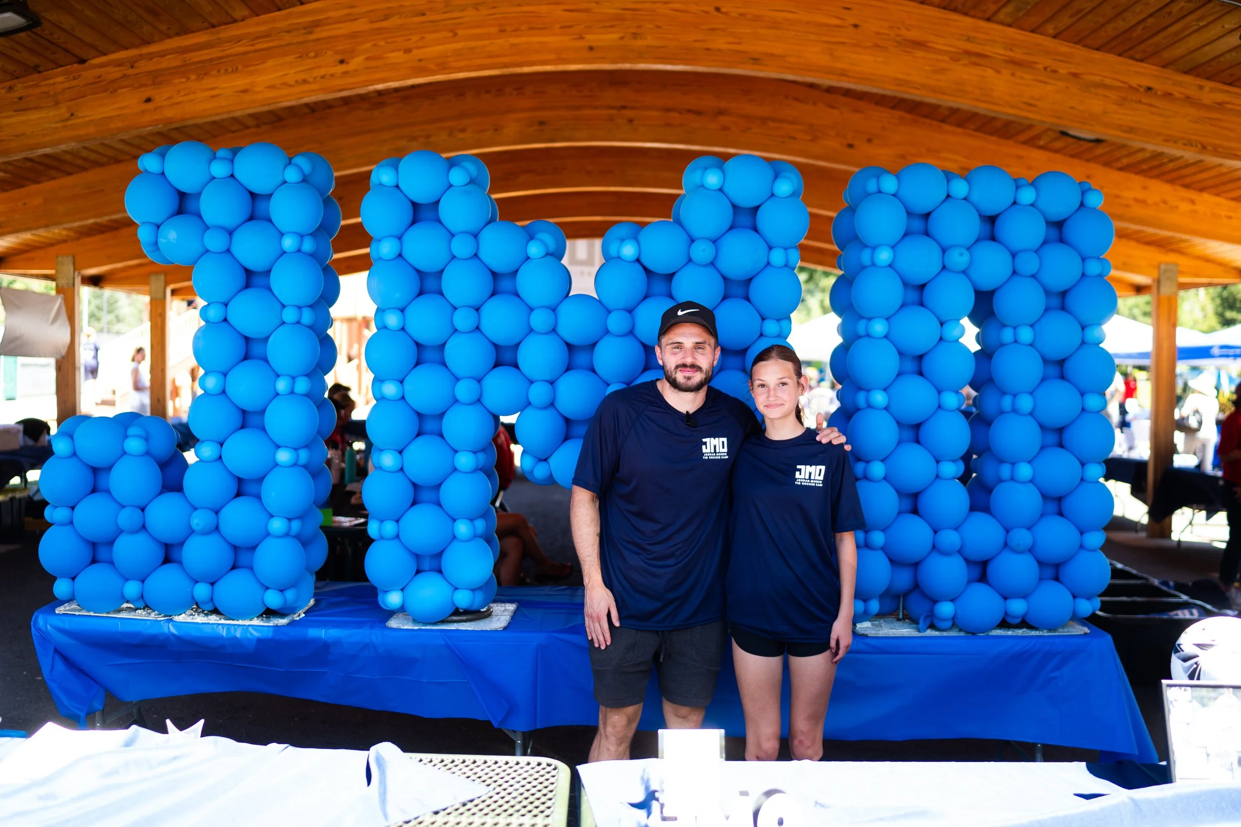 Two people, a man and a girl, standing in front of a blue balloon arch at an outdoor event under a wooden pavilion.