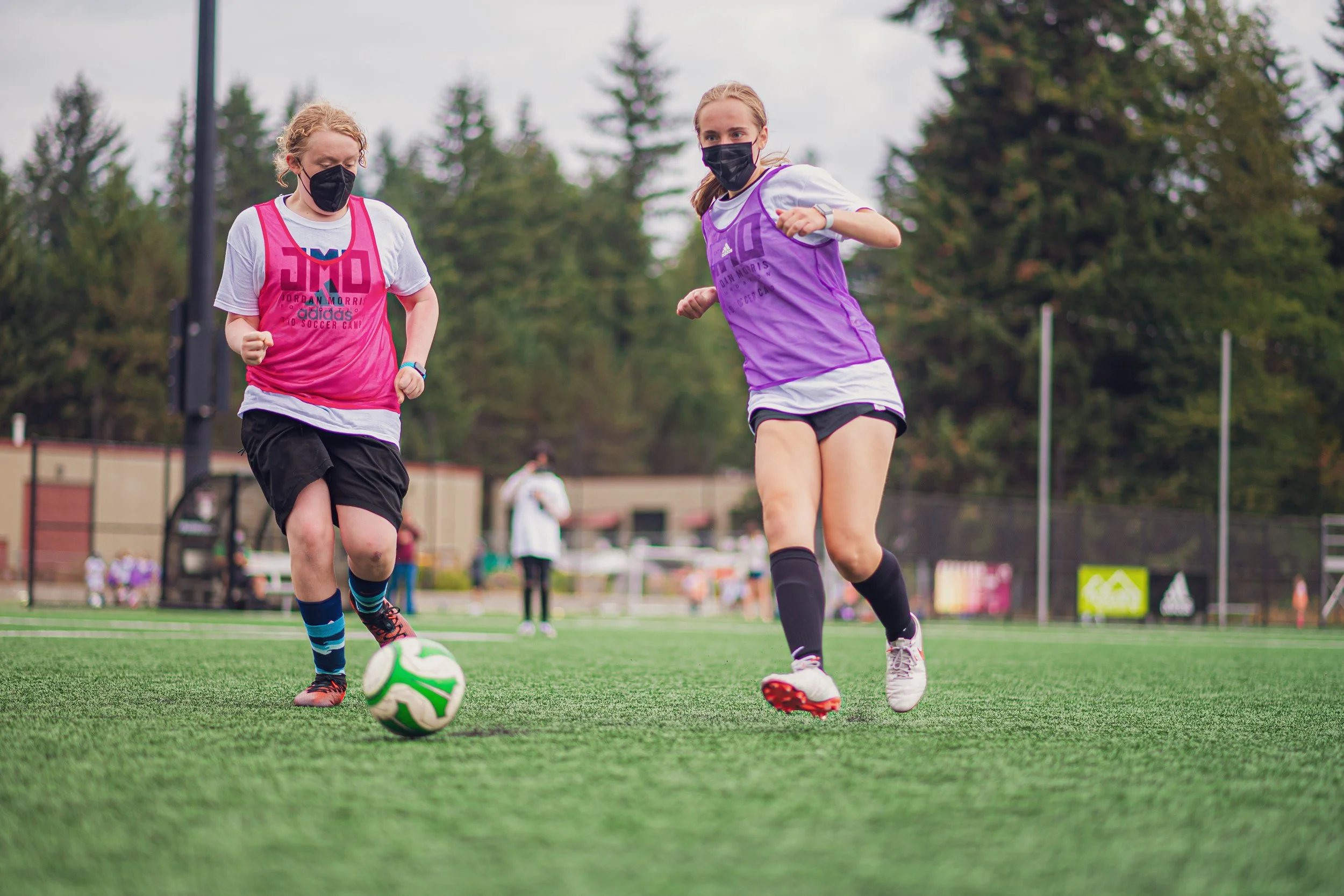 Two young women, wearing masks, playing soccer on a field with trees in the background. One woman in a pink vest is kicking a green and white soccer ball while the other in a purple vest runs alongside her.
