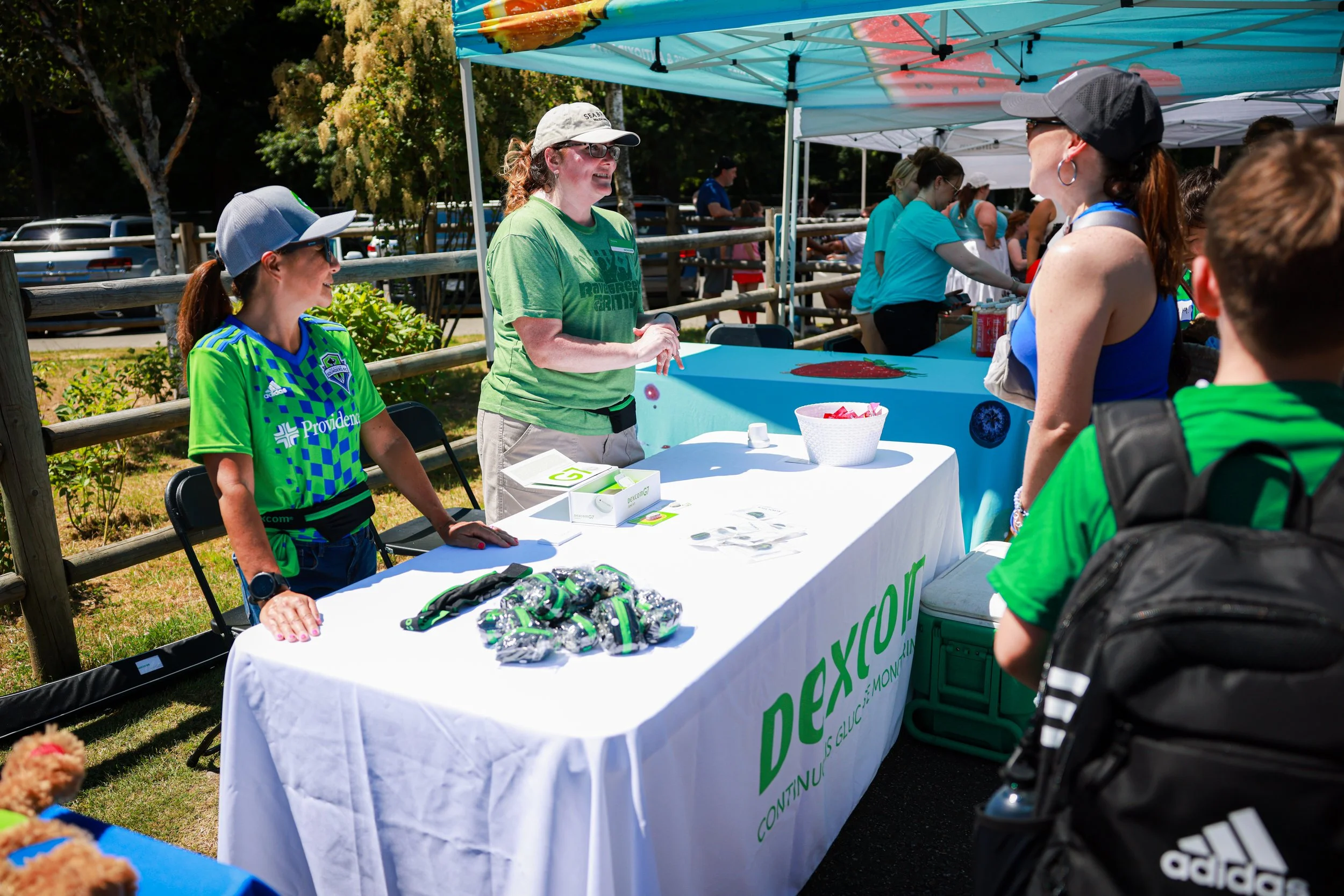 At an outdoor event, a woman in a green T-shirt and glasses is talking to a group of people at a booth with a blue tablecloth, under a canopy. The booth displays promotional items including wristbands and snacks, and has a sign with the word 'dexcom'