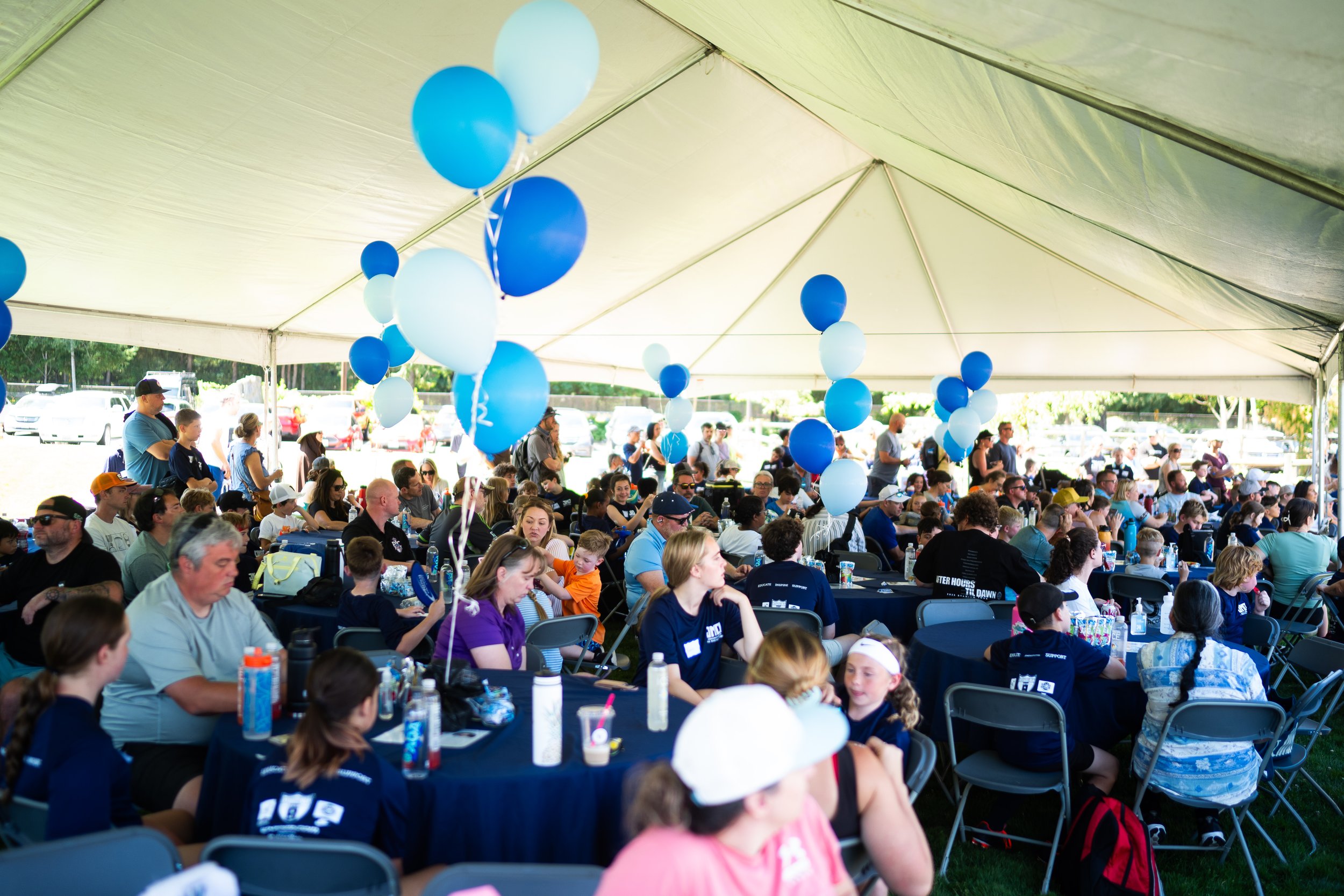 A large gathering of people sitting at tables under a white event tent, decorated with blue and white balloons, during a daytime outdoor event.