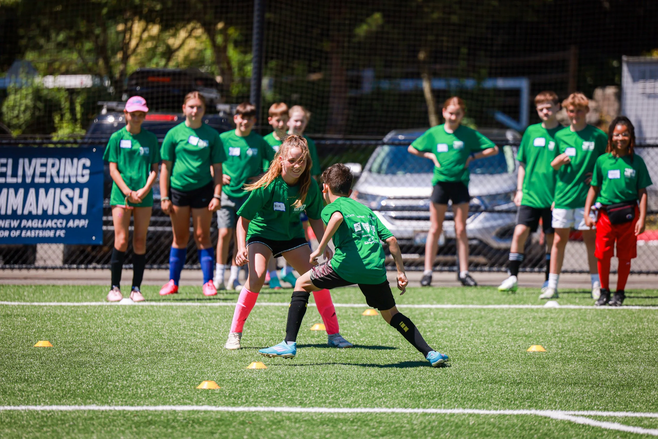 Children participating in a soccer drill on a green field, with a woman guiding them, surrounded by other kids in green shirts and a chain-link fence in the background.