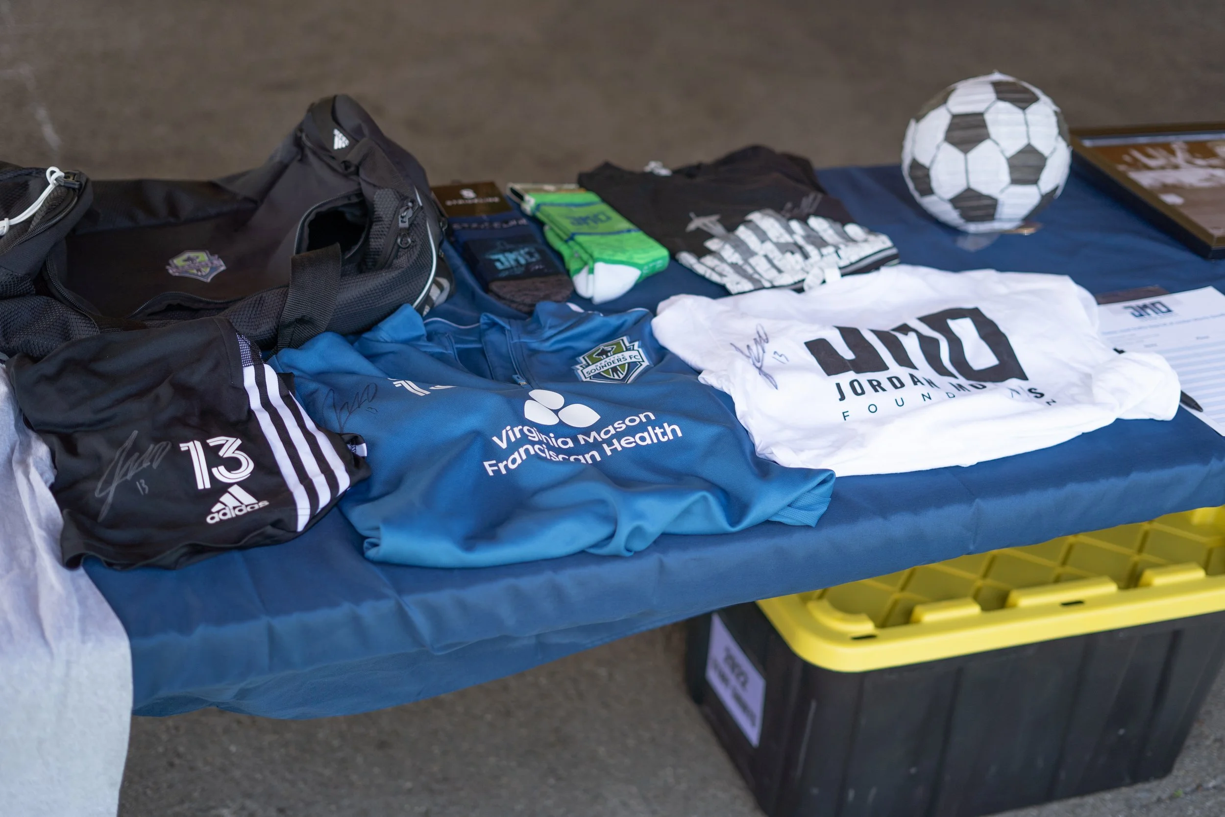 Table with sports clothing and accessories, including a soccer ball, signed team jerseys, socks, and bags, on display at an event.