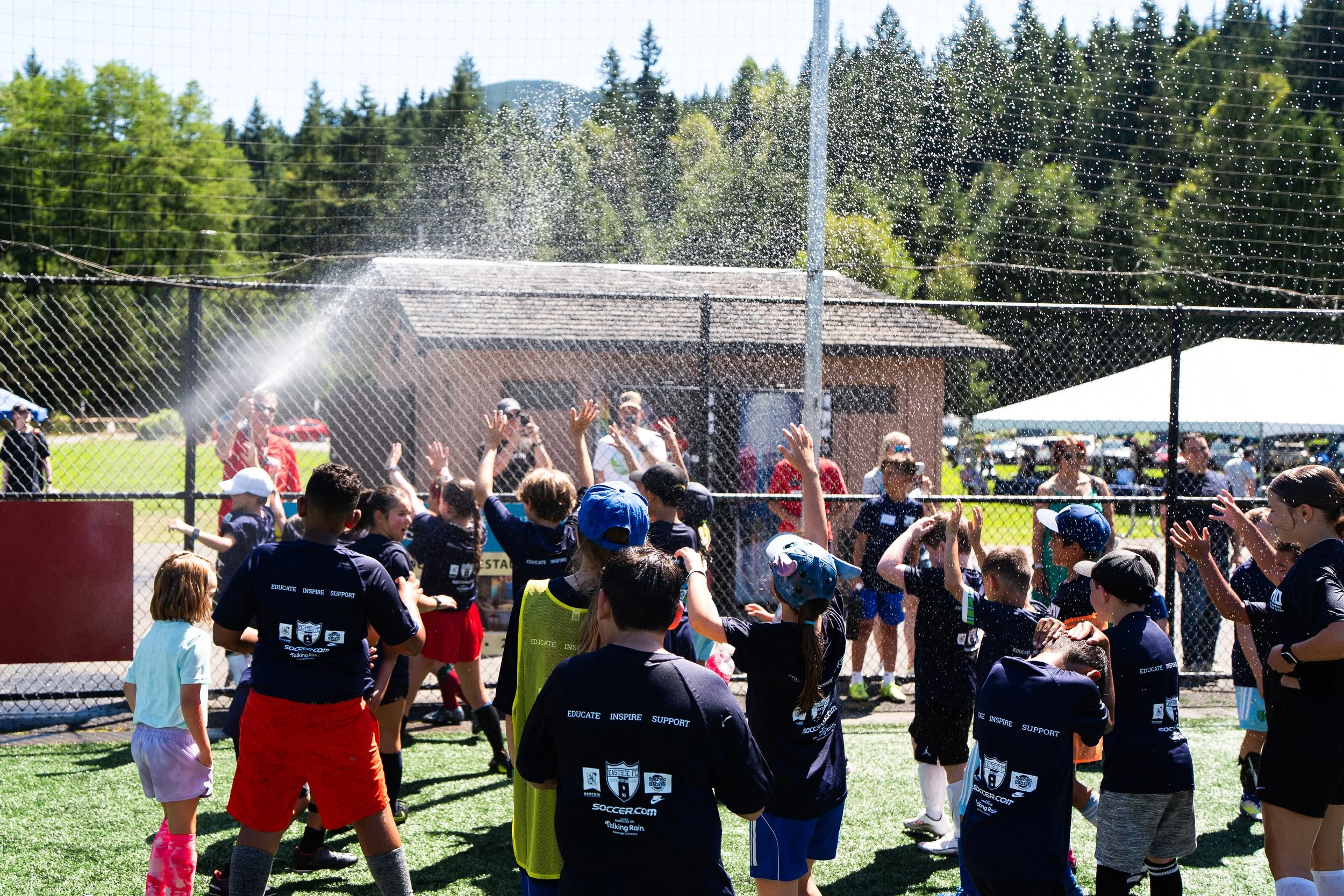 A group of children and adults at a sports field, with some raising their hands under water being sprayed from a sprinkler, surrounded by a chain-link fence, with trees and a blue sky in the background.