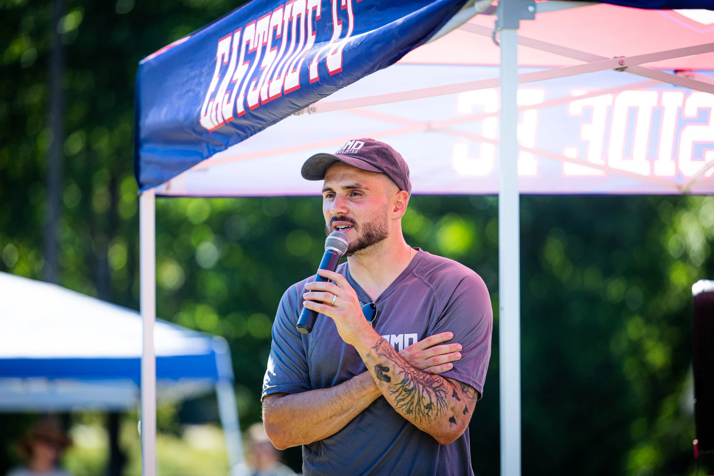 A man with tattoos on his arms, wearing a gray t-shirt and baseball cap, speaking into a microphone at an outdoor event, with a blue canopy overhead and green trees in the background.