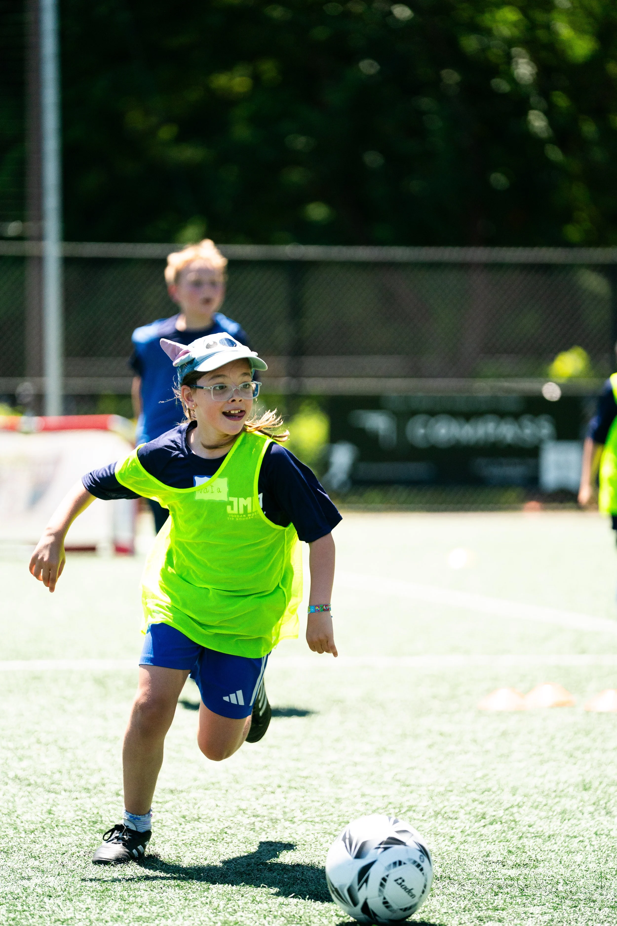A young girl wearing glasses, a white hat with animal ears, a yellow vest, and blue shorts is running on a grassy field towards a soccer ball during a sunny day. In the background, there are other children, trees, and a chain-link fence.