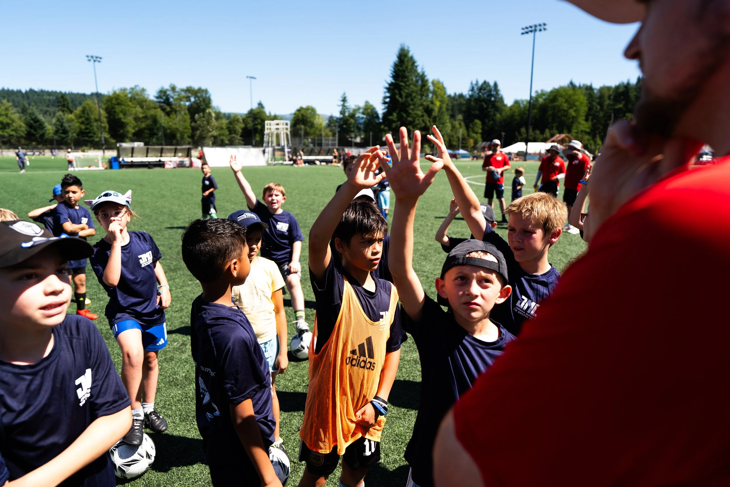 Group of children on a soccer field, some raising their hands, with a coach or adult figure in red talking to them during a sunny outdoor soccer practice or game.
