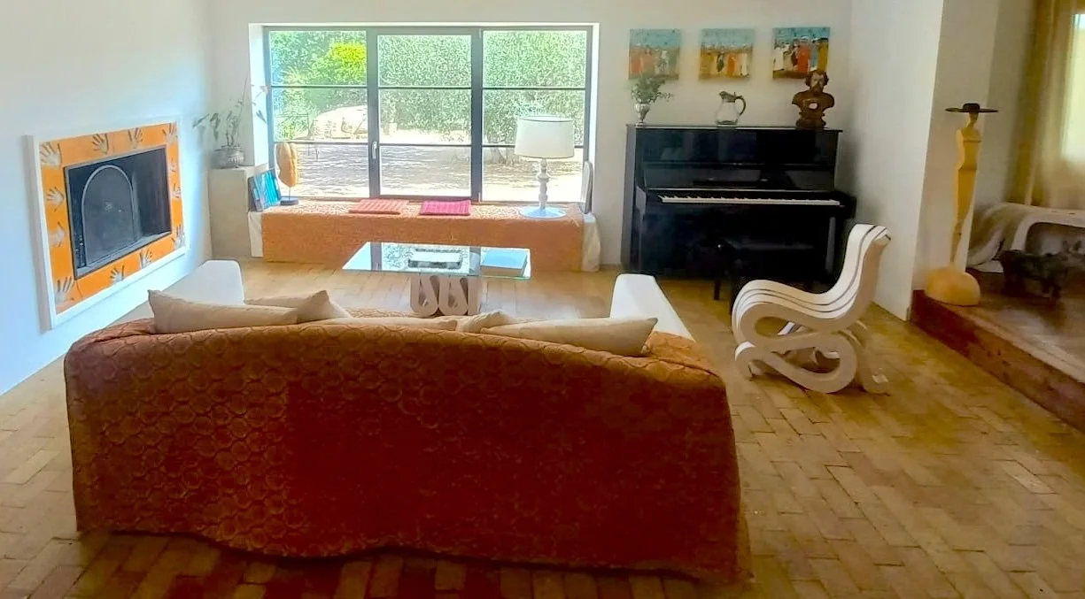 Salotto con divano marrone, poltrona bianca, piano nero, finestra con vista su giardino, decorazioni colorate alle pareti.