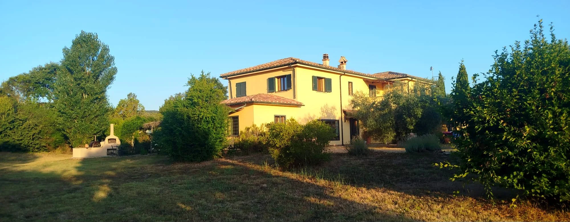 Casa rustica in campagna con giardino verde e alberi, sotto un cielo azzurro.