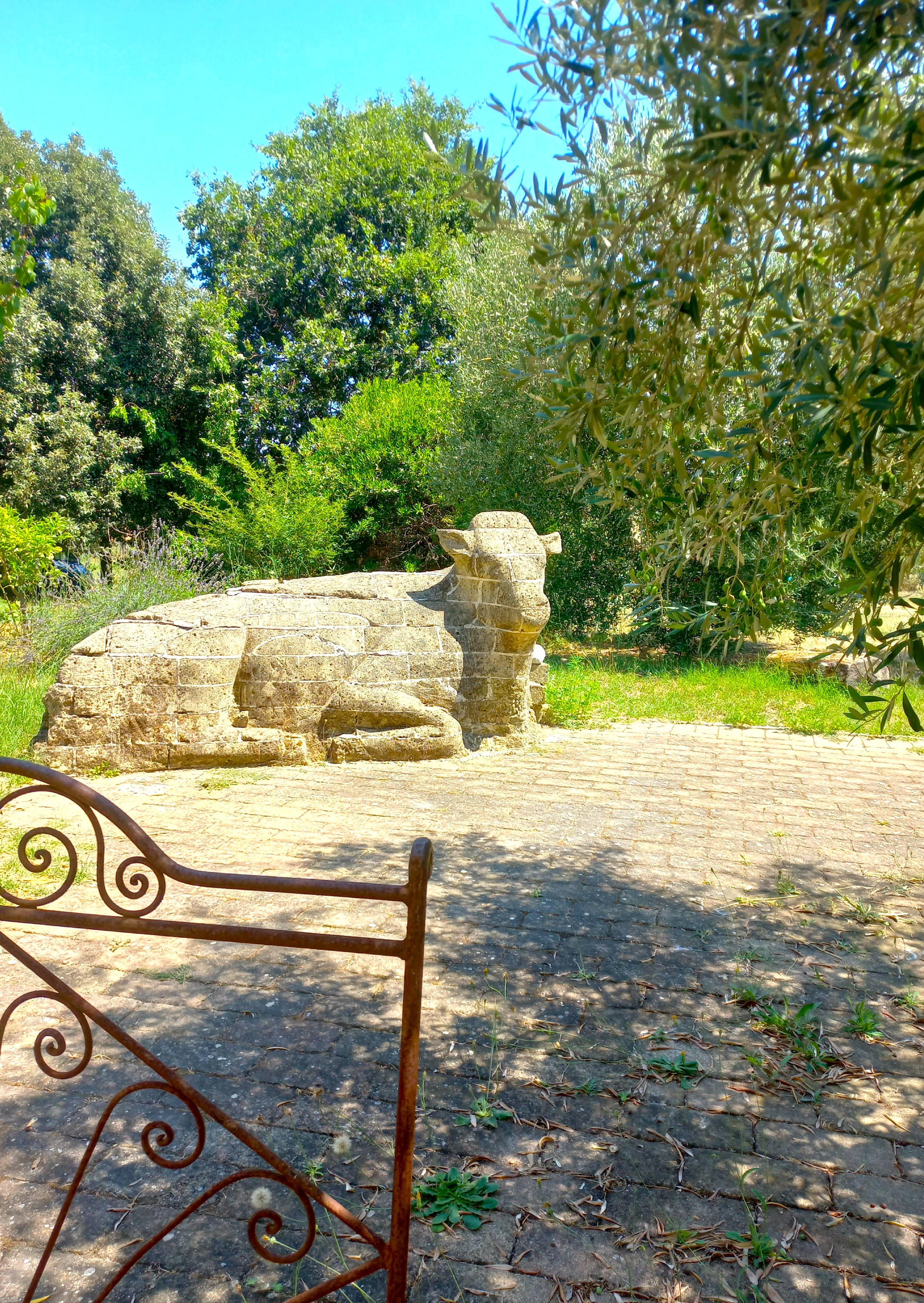 Scultura di una mucca in pietra di tufo  in un giardino con piante verdi e cielo azzurro.