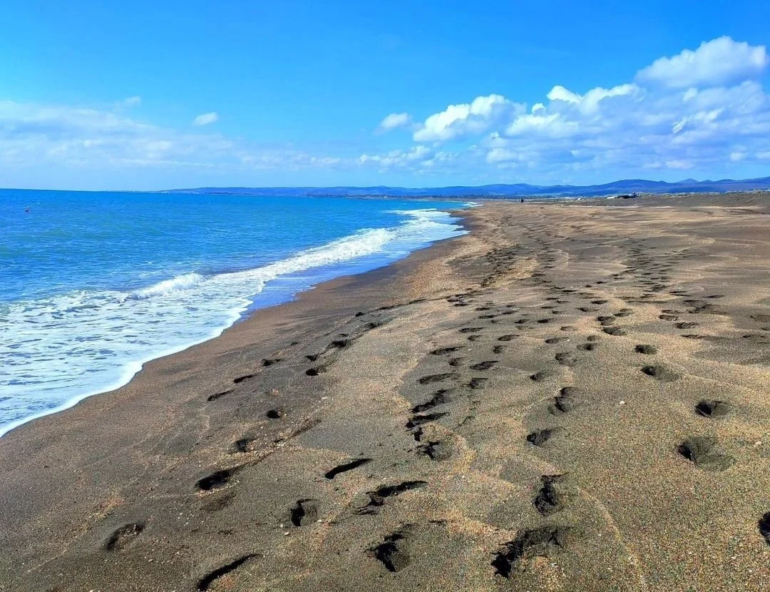 Spiaggia sabbiosa vista dall'orizzonte, con impronte sulla sabbia e onde che si infrangono sulla riva, sotto un cielo azzurro con nuvole sparse.