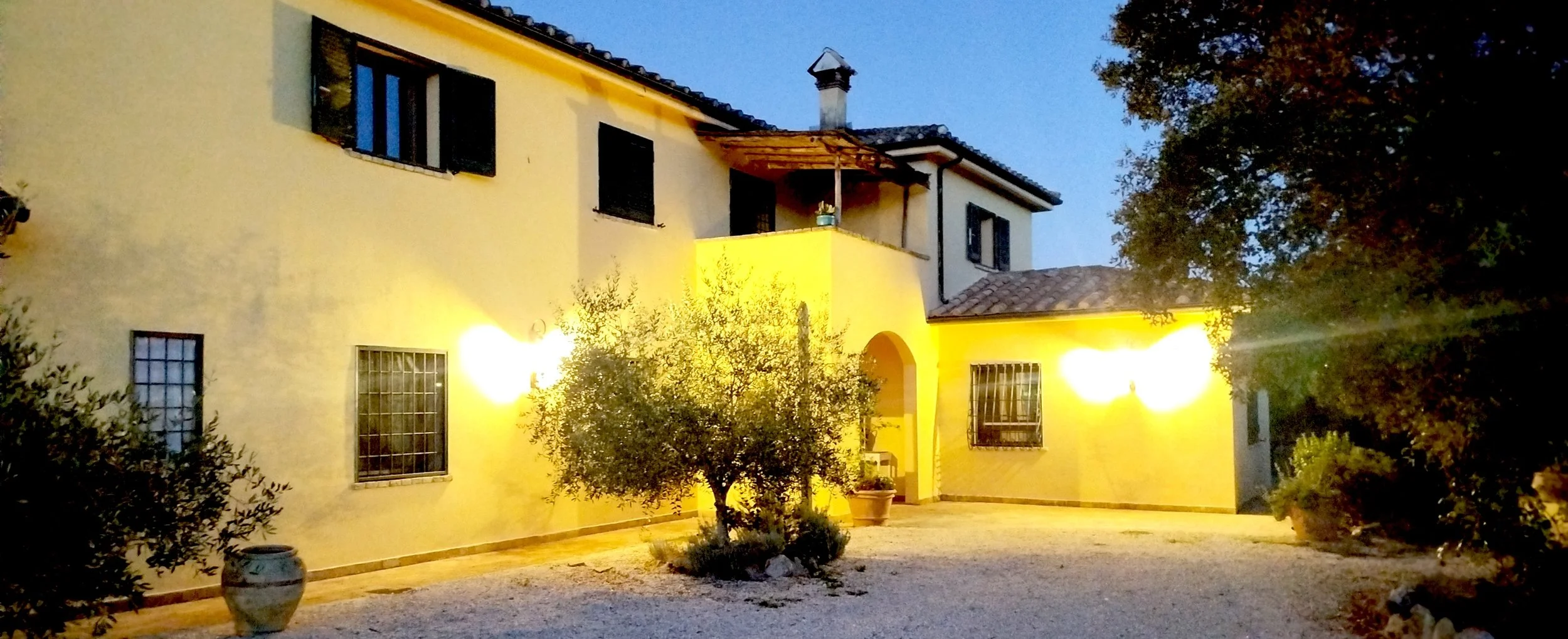 Casa con pareti gialle e luci esterne accese, circondata da alberi e piante, al tramonto o in prima sera.