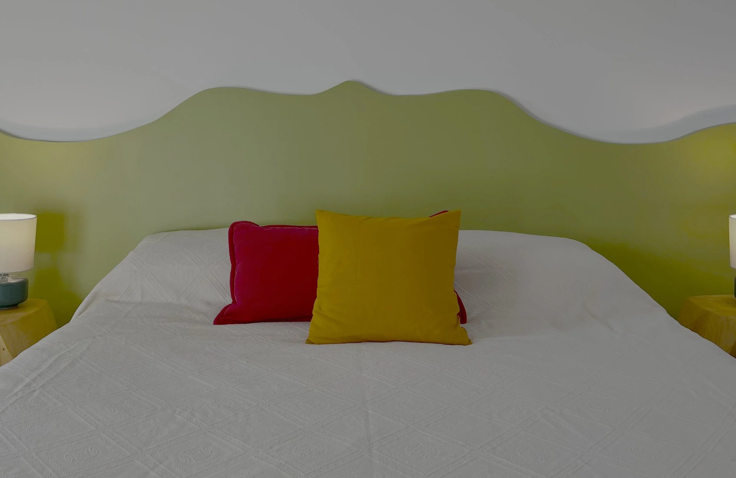 Letto con cuscini arancione e rosso, testata verde ondulata, lampada a sinistra, tavolino di legno a destra.