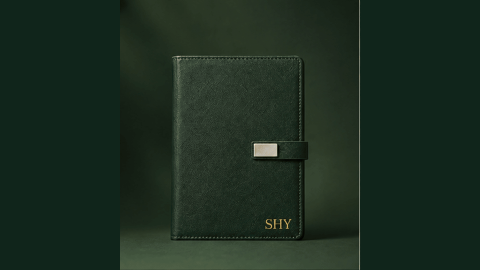 A closed dark green planner or journal with a strap and a metallic rectangular detail, with the word "SHY" embossed in gold at the bottom right corner.
