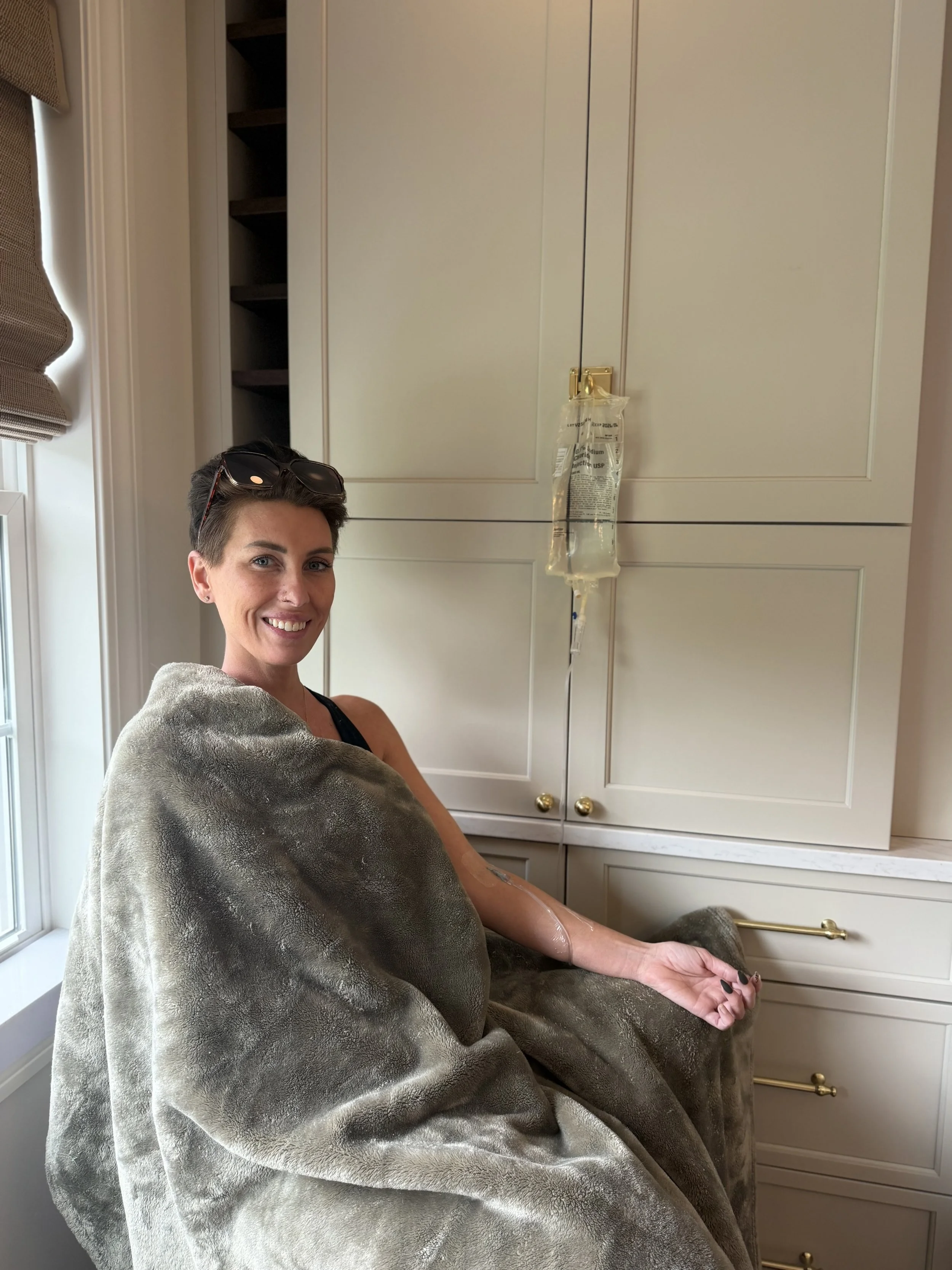 A woman in a hospital bed with an IV drip attached to her arm, sitting in a room with white cabinetry and window with blinds, smiling at the camera.