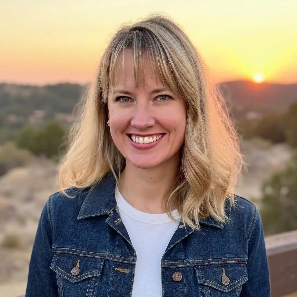 A woman with blonde hair smiling outdoors during sunset, wearing a denim jacket and white shirt, with a scenic landscape in the background.