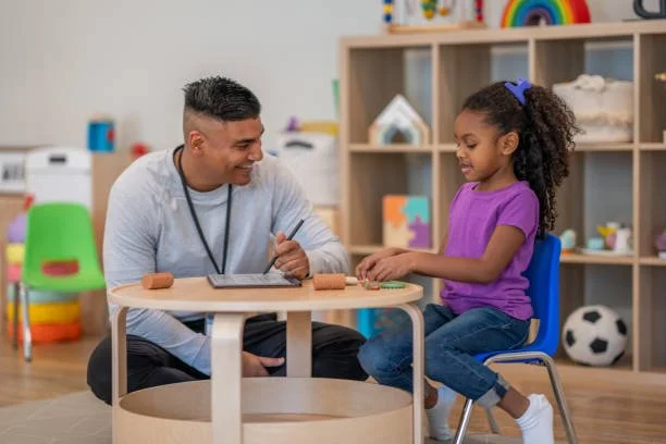 A young girl in a purple shirt sitting at a table with a therapist in a play therapy room with toys and a bookshelf in the background.