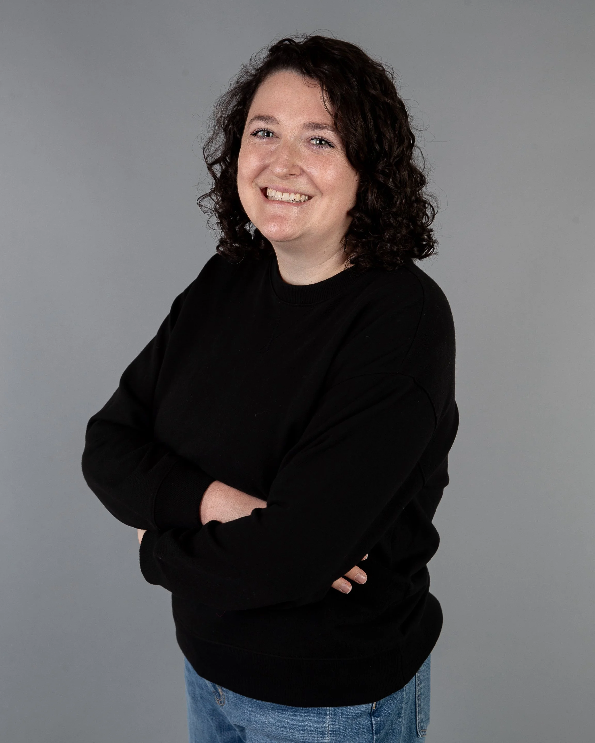 A person with curly dark hair wearing a black sweater and light blue jeans, smiling against a plain gray background.