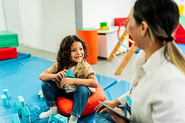 A young girl sitting on an orange cushion in a therapy or playroom, smiling and talking to a woman who appears to be a therapist or counselor, with colorful toys and play equipment in the background.