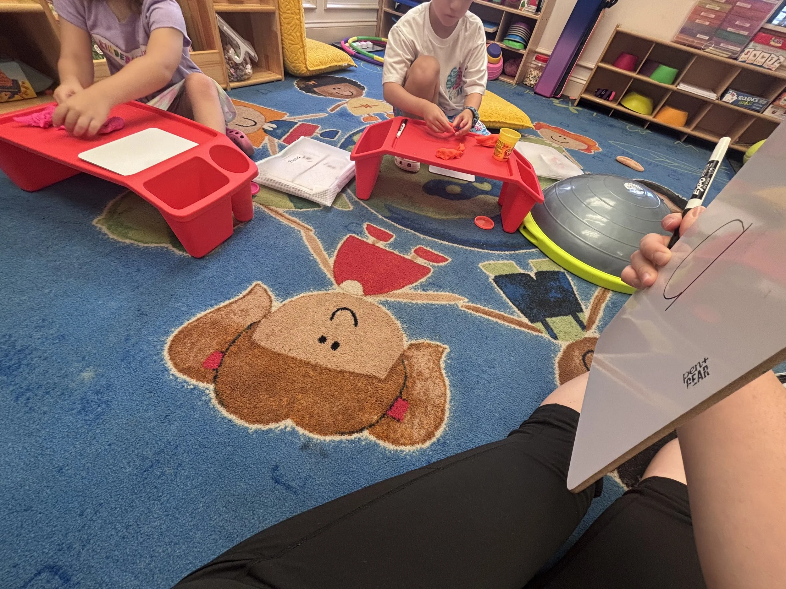 Children sitting on a colorful learning rug, engaging in arts and crafts activities at a classroom or daycare setting, with shelves of toys and supplies in the background.