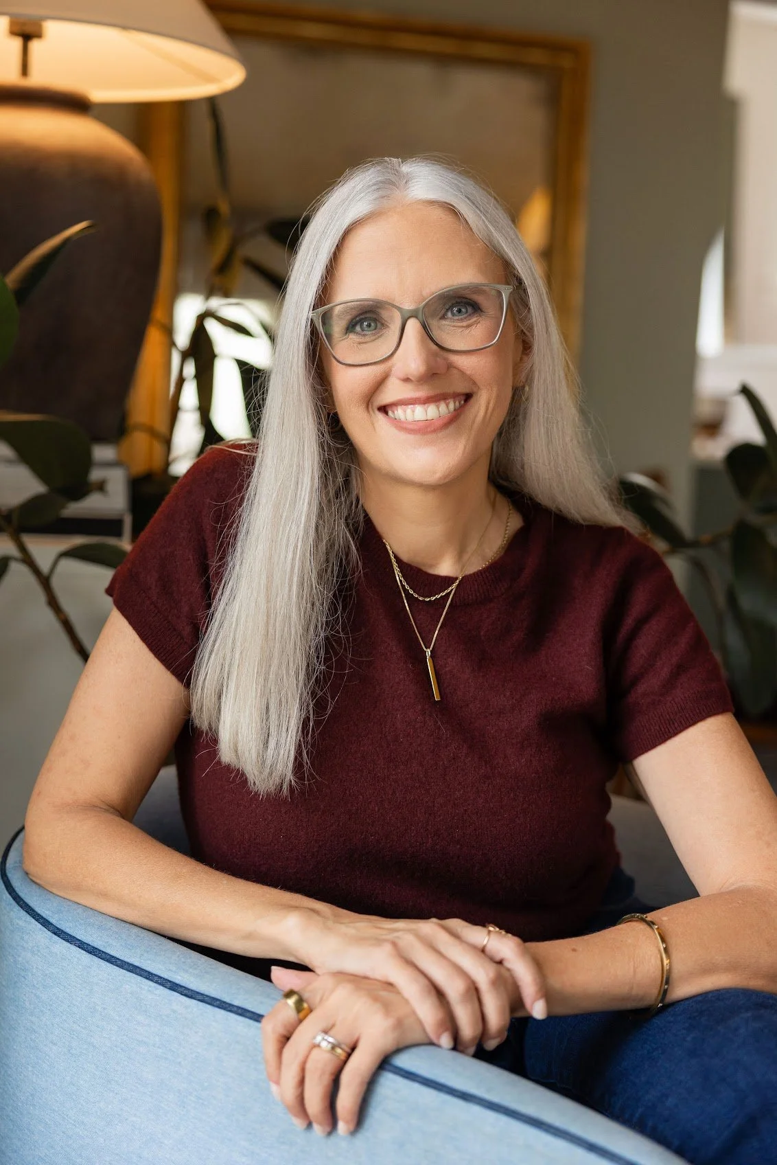 A woman with long gray hair and glasses smiling, sitting on a light blue chair indoors, wearing a maroon shirt and layered necklaces.