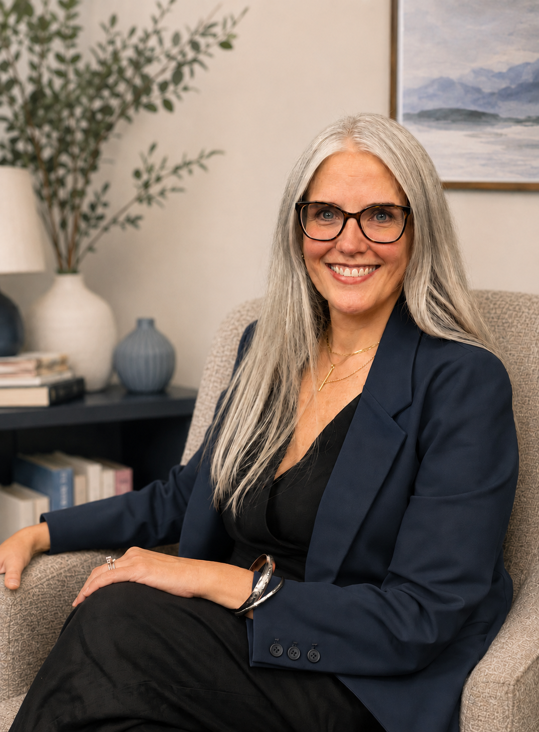 A smiling woman with long gray hair, wearing glasses, a navy blazer, and a black top, sitting in a beige armchair in an office or living room. There are decorative vases, books, and a painting of mountains and a lake in the background.