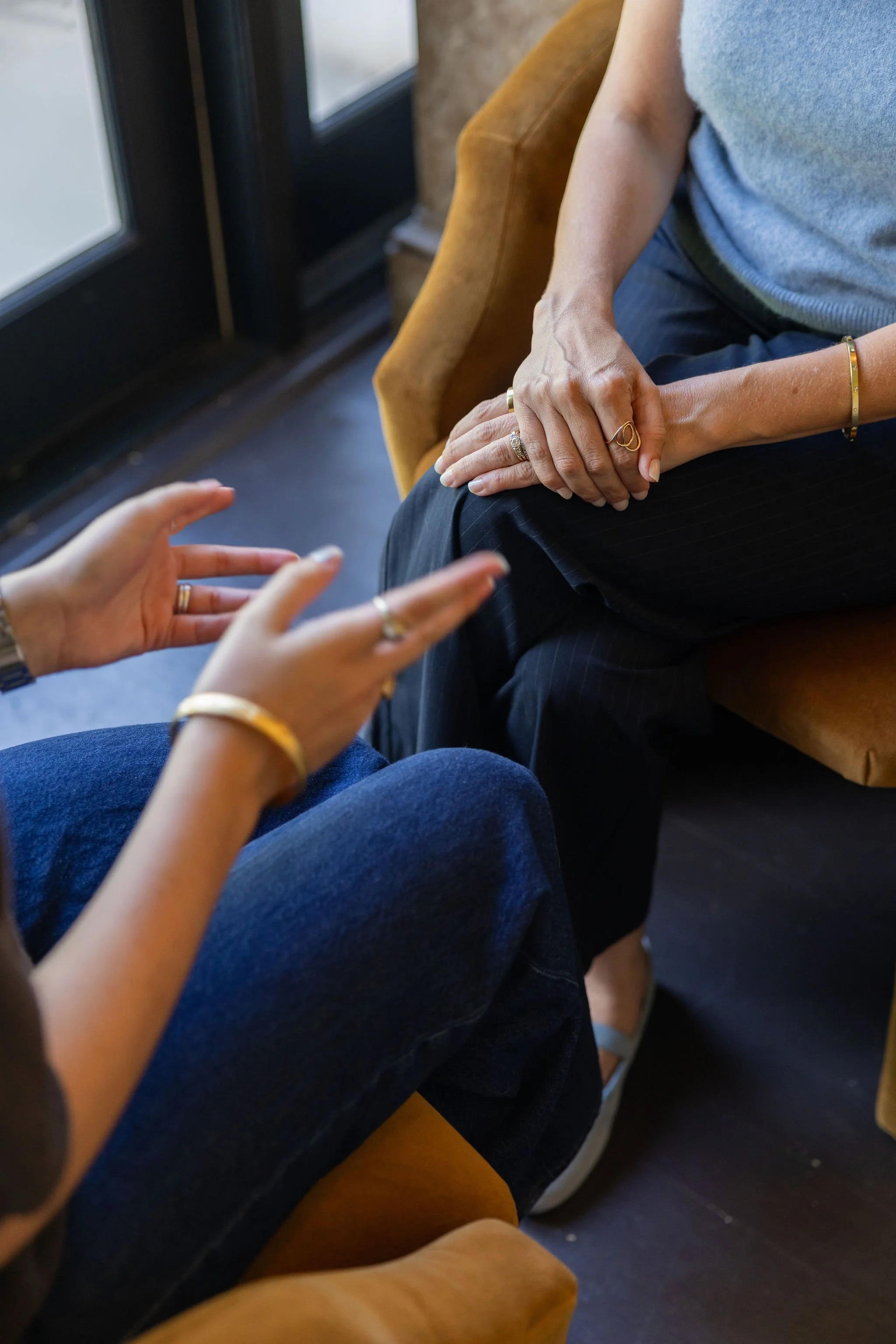 Close-up of two people holding hands, one wearing rings and a bracelet, sitting on a tan chair near a window.
