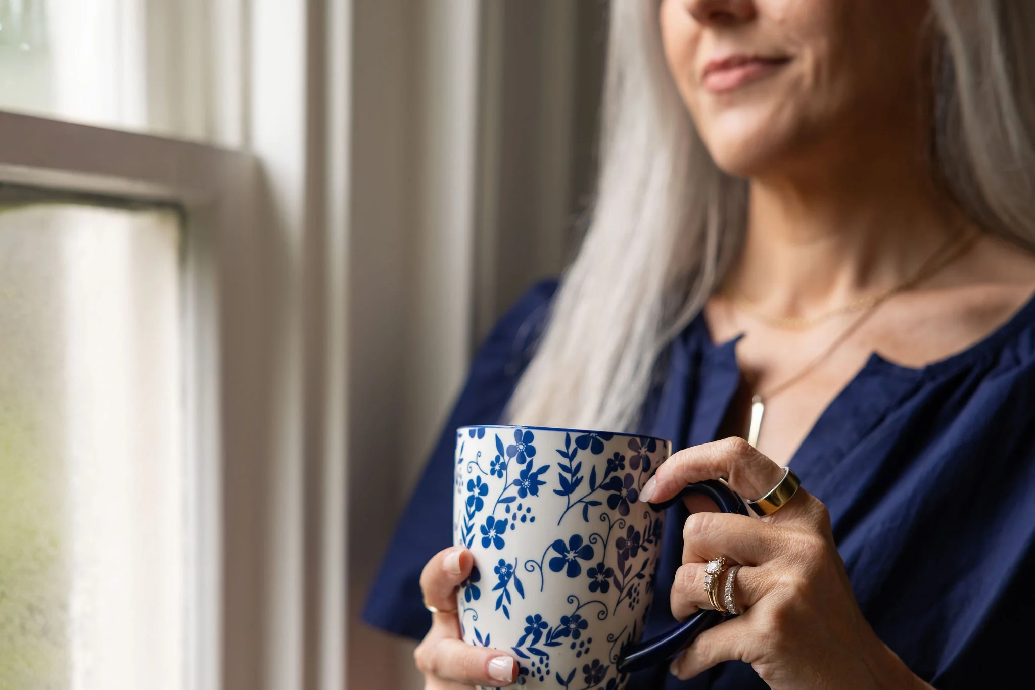 A woman holding a blue and white floral mug, wearing rings and a gold necklace, with her face partially visible, near a window.