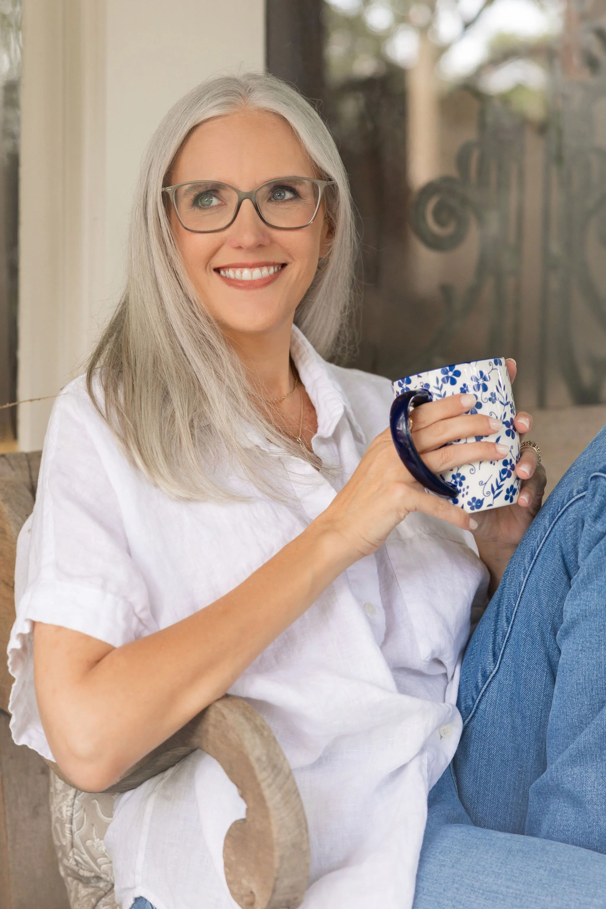 Smiling woman with gray hair and glasses, holding a patterned mug, sitting outdoors by a window with a blurred garden view behind her.