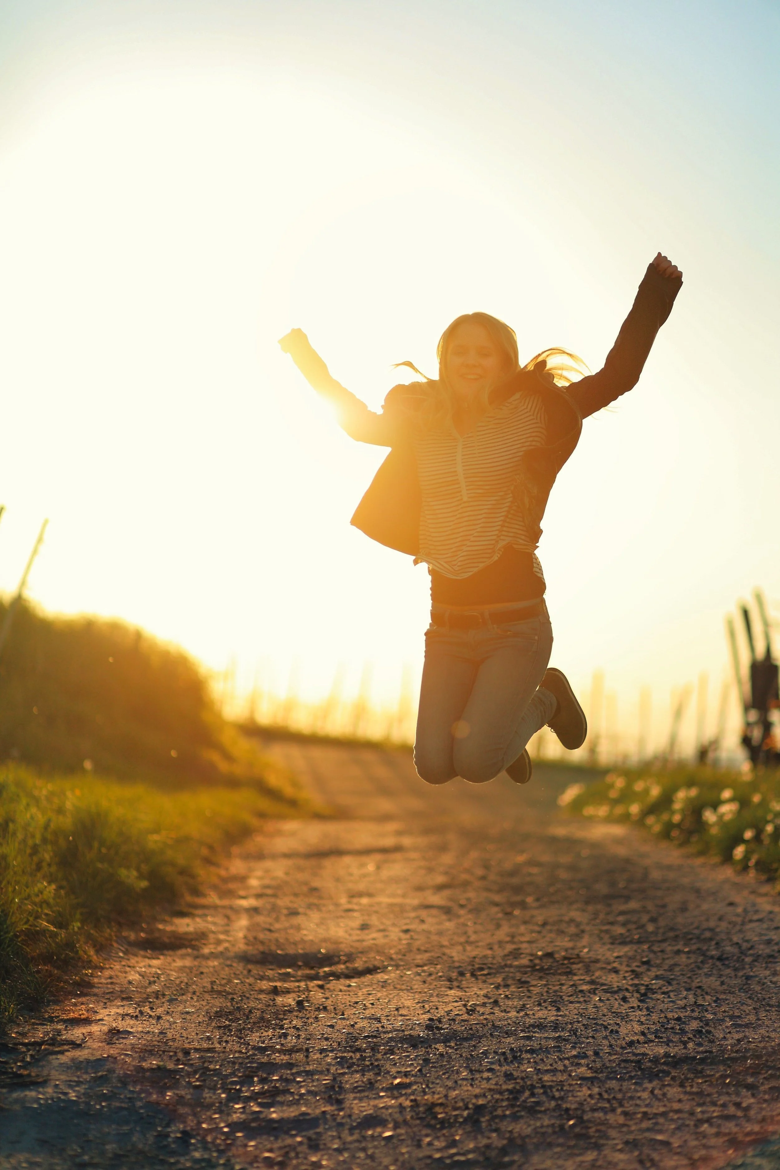 A woman jumping in the air with her arms raised during sunset on a dirt path.