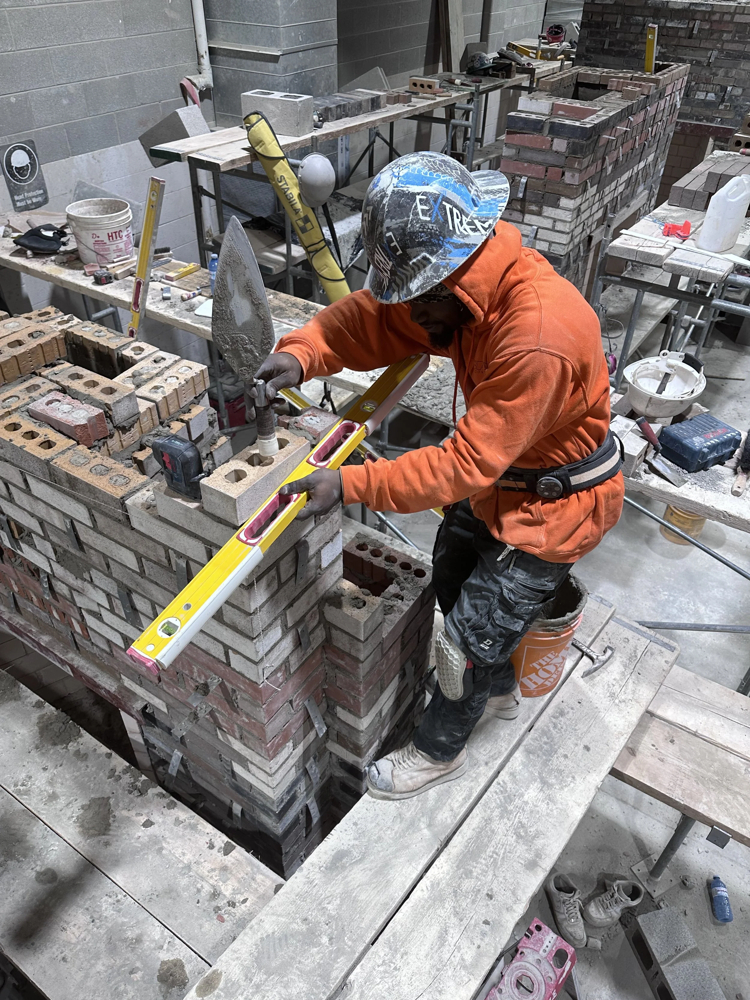 A construction worker in an orange hoodie and camouflage pants is working on a brick wall at a construction site. He is using a level tool to ensure proper alignment of bricks. The site has various construction tools, materials, and scaffolding around him.