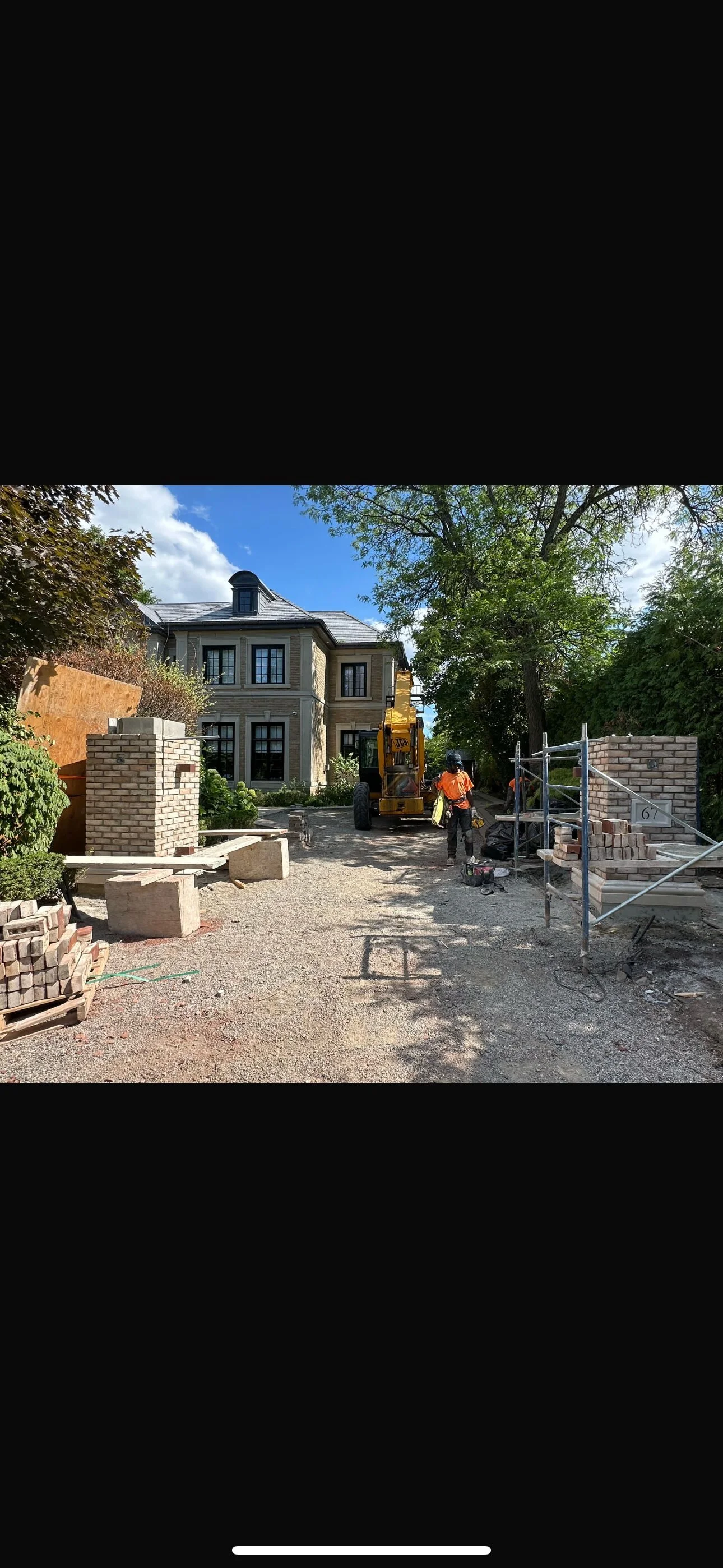 Construction workers and equipment on a residential driveway, with a large house in the background and brick walls under construction.