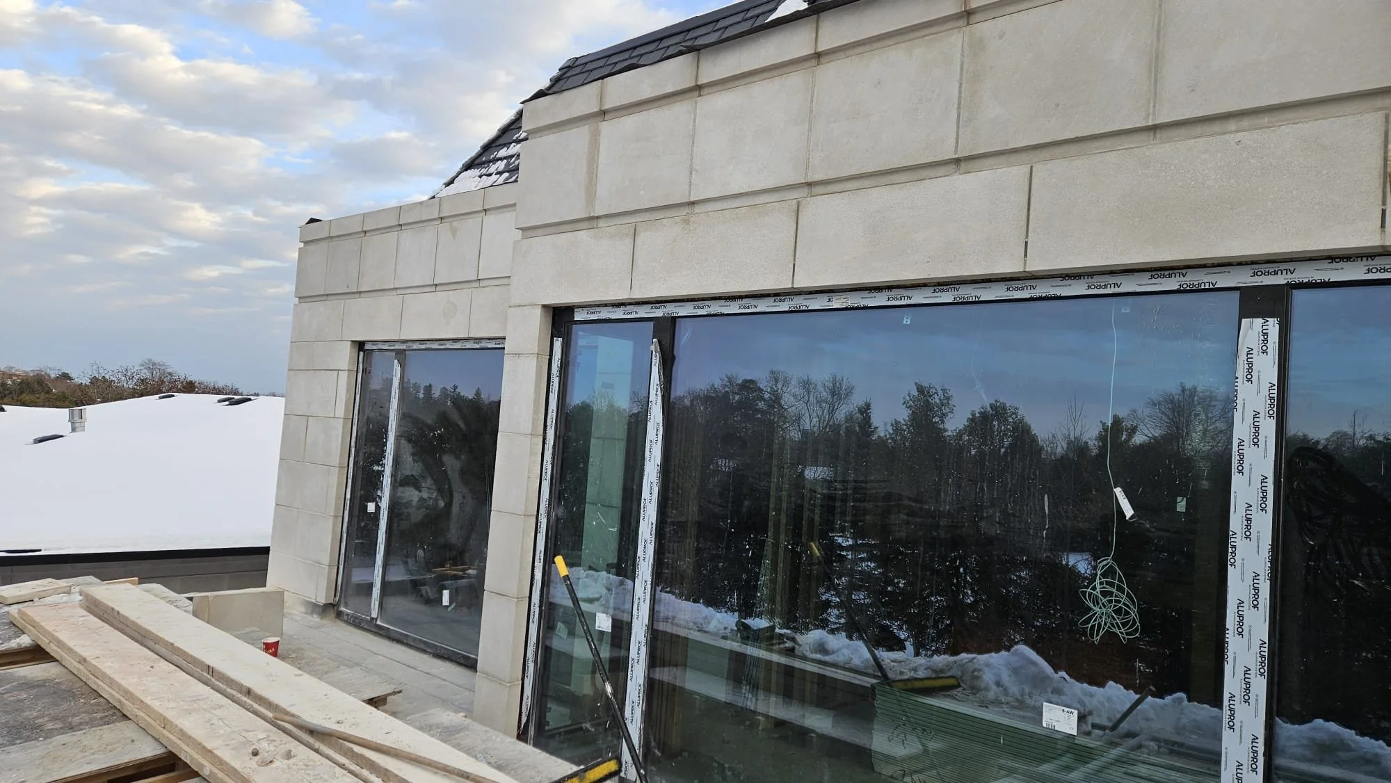 Construction site of a house with large glass windows and stone exterior walls, snow on the ground, and trees in the background under a partly cloudy sky.