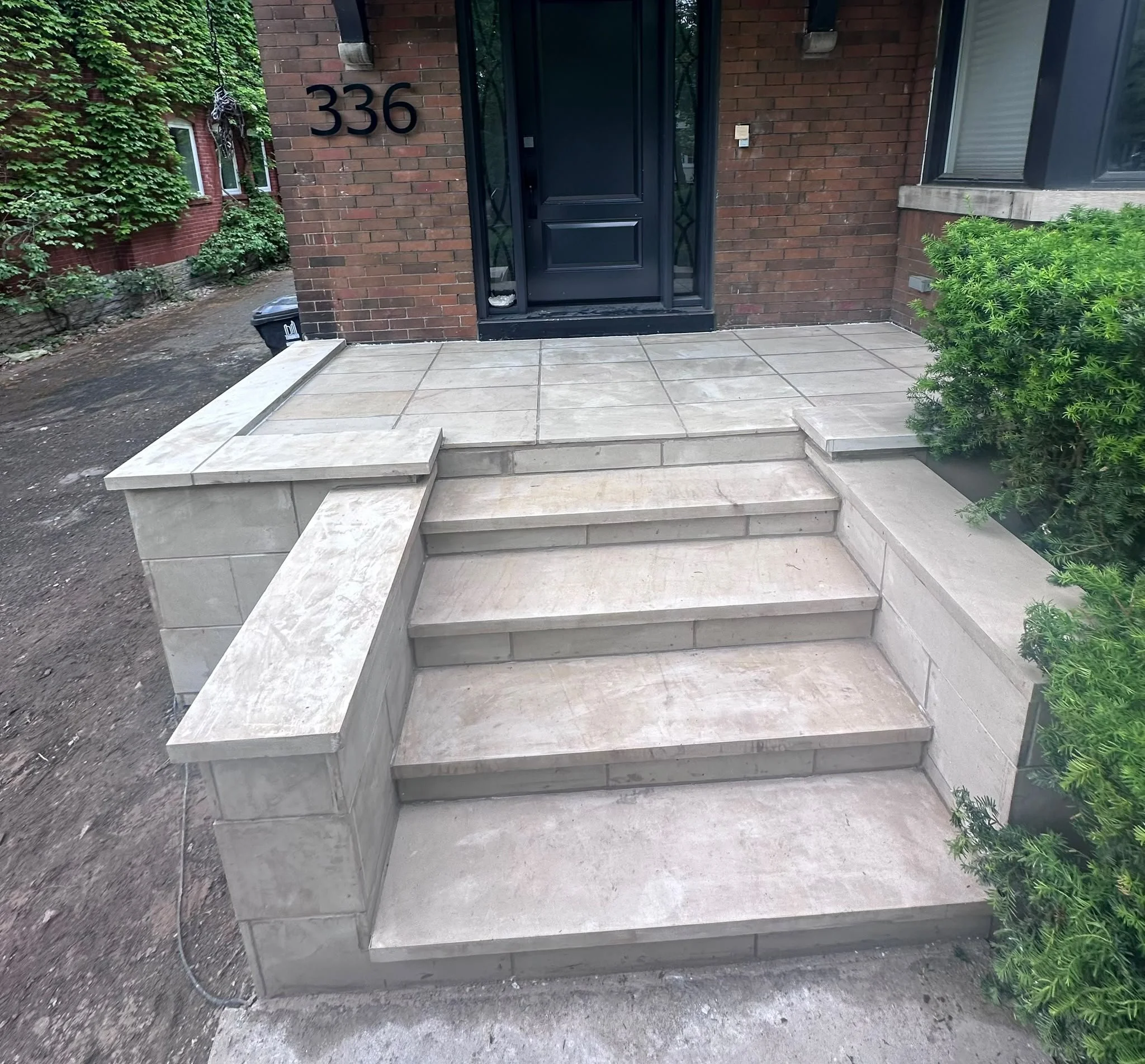 Concrete steps leading up to a black front door with a security screen, the house number 336 on a brick wall, green shrubs on the right, and a walkway with dirt and a trash bin on the left.