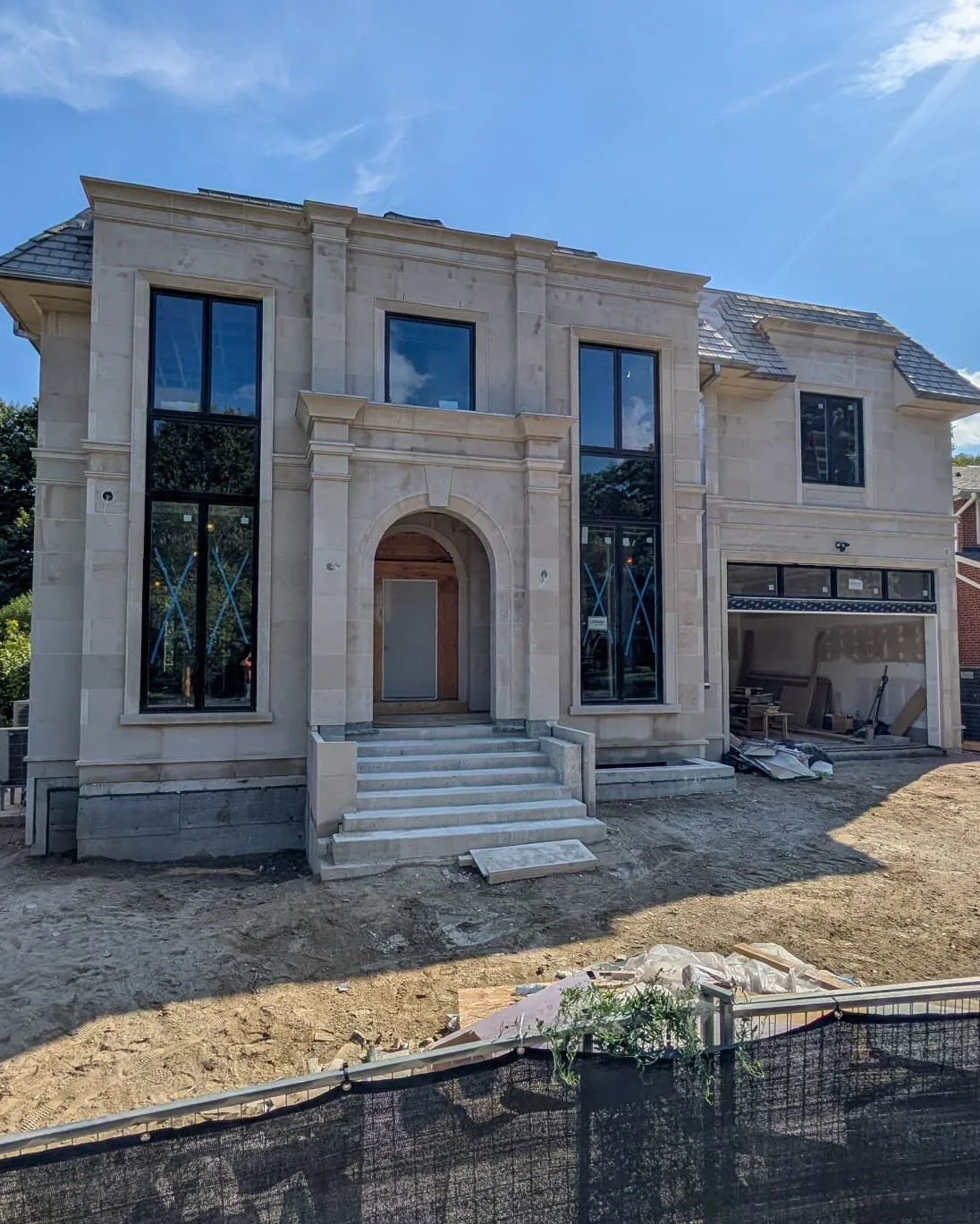 Under construction two-story house with stone exterior, large windows, arched front entrance, and stairs leading up to the door.