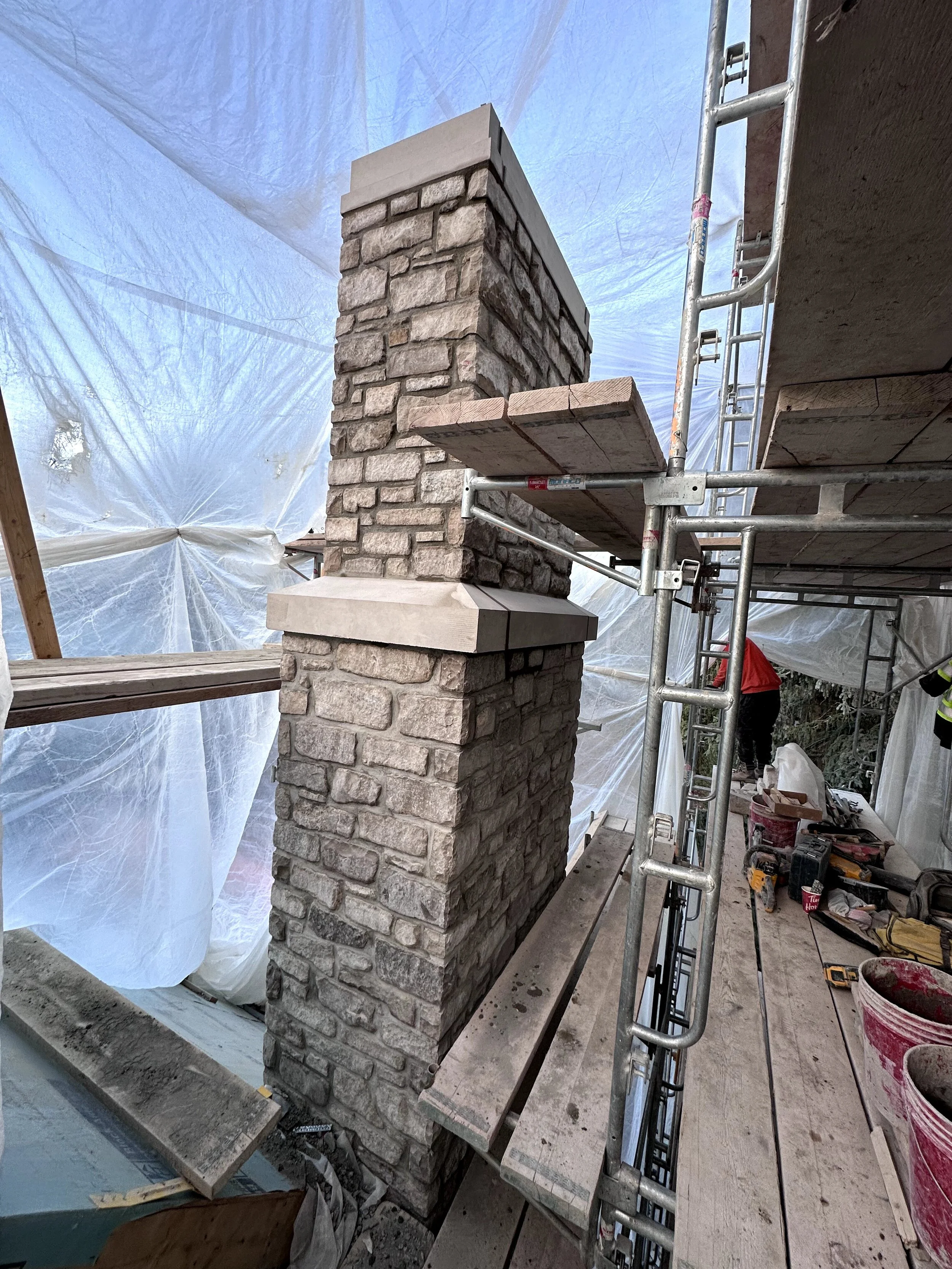 Construction site with a stone chimney being built, scaffolding and a worker in the background, tools and materials scattered around.