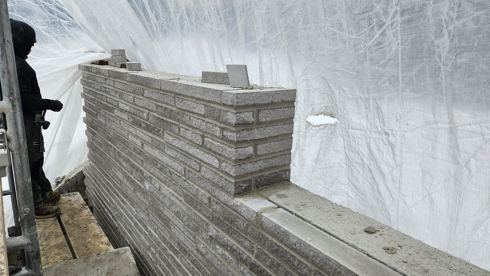 A worker standing next to a partially built brick wall inside a construction site with white protective sheeting.